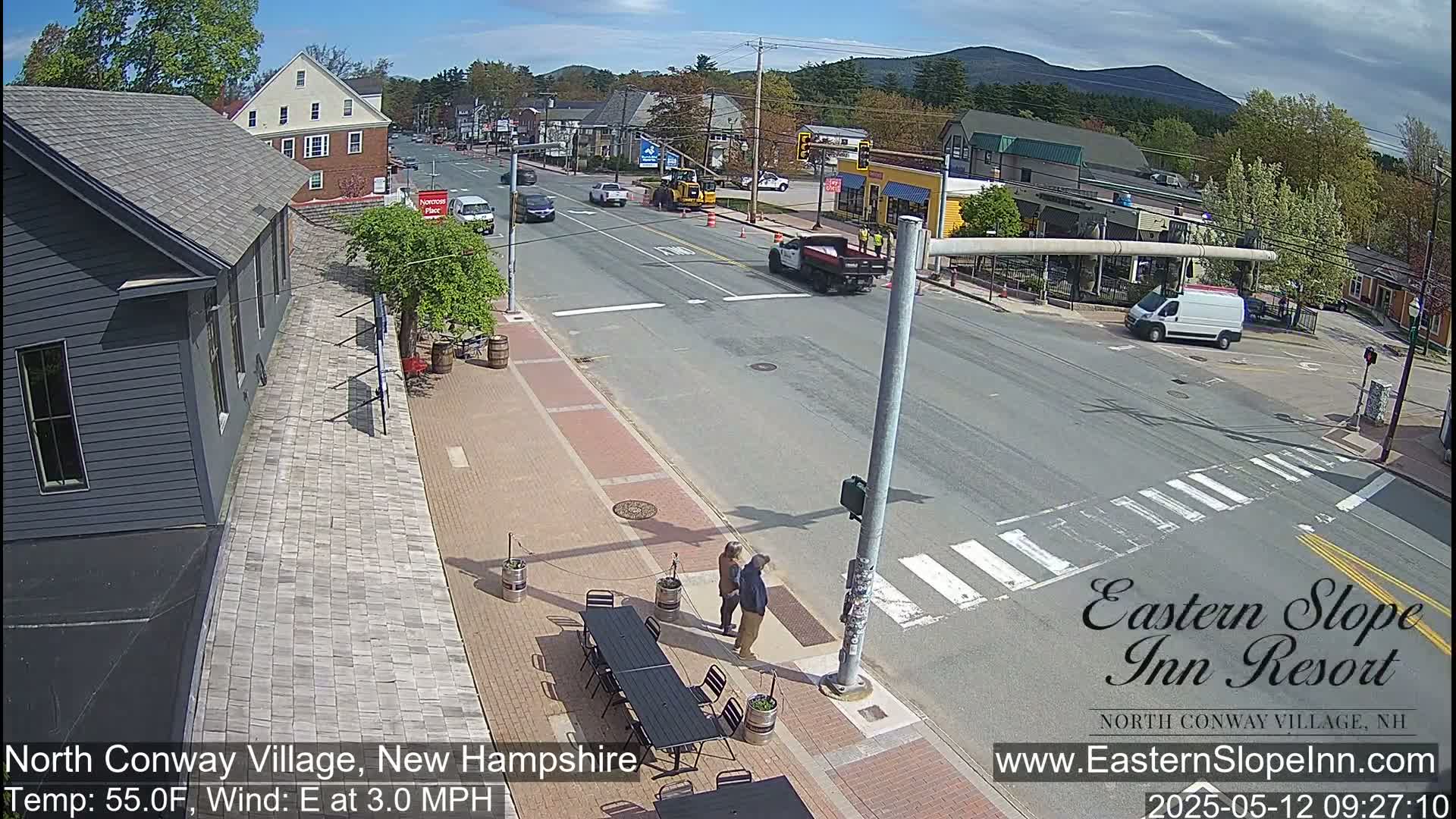 A street scene on a partly sunny day shows a village intersection with cars and pedestrians, and a building with outdoor seating.