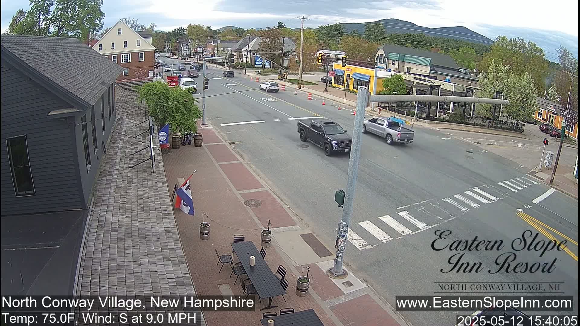 A partly cloudy day shows a street scene with several vehicles and pedestrians, outdoor seating at a restaurant, and buildings in a village setting.