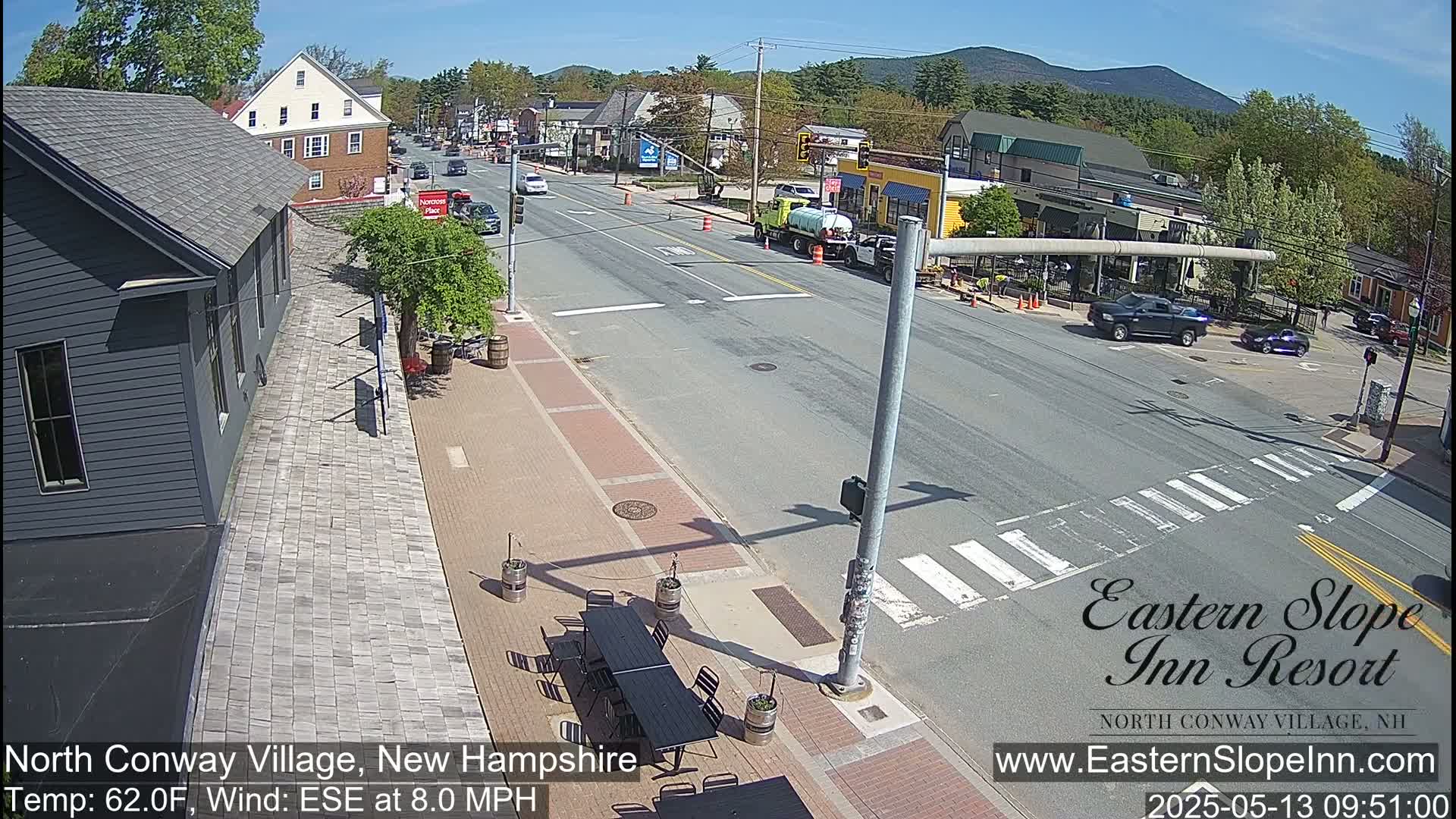 A sunny street scene shows a mostly empty road with several cars, buildings lining the street, and outdoor seating at a patio cafe.