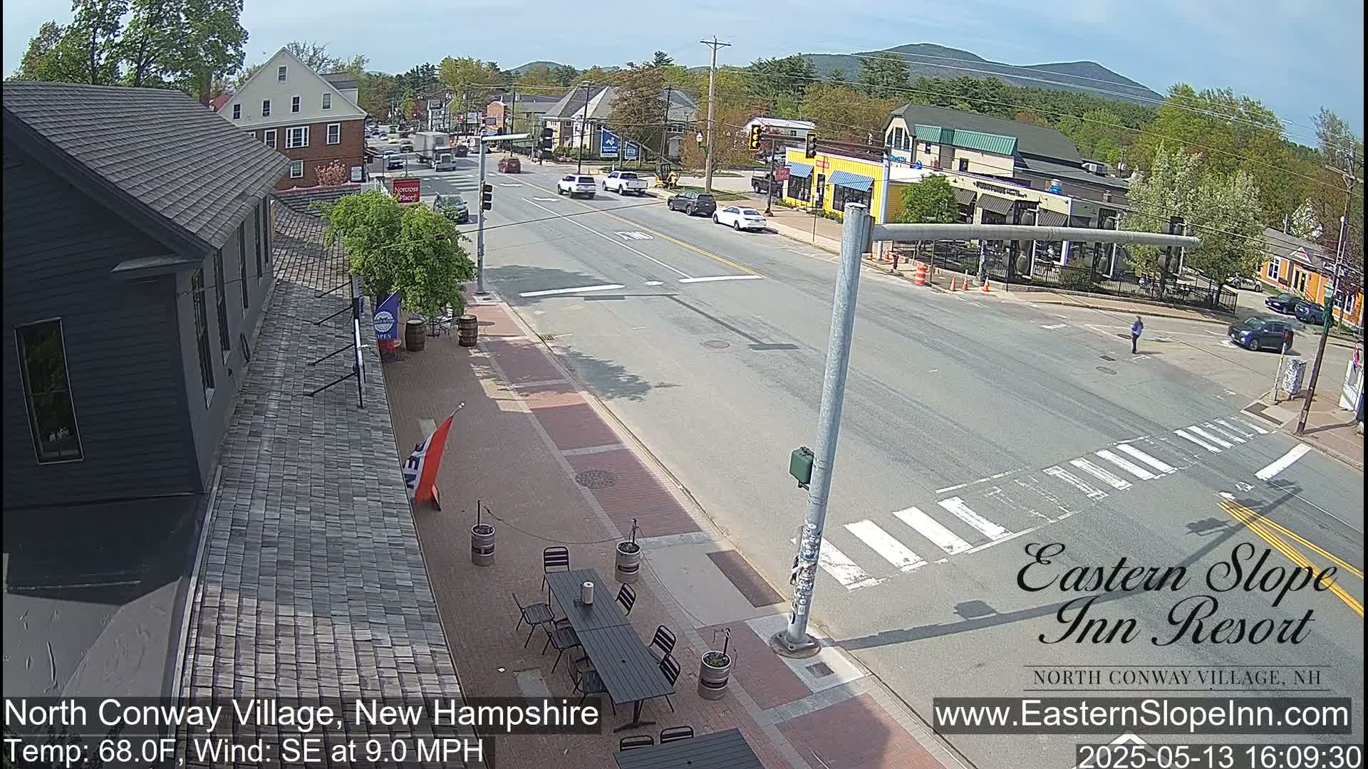 A sunny view of a town street scene with several cars, pedestrians, and outdoor seating at a restaurant.