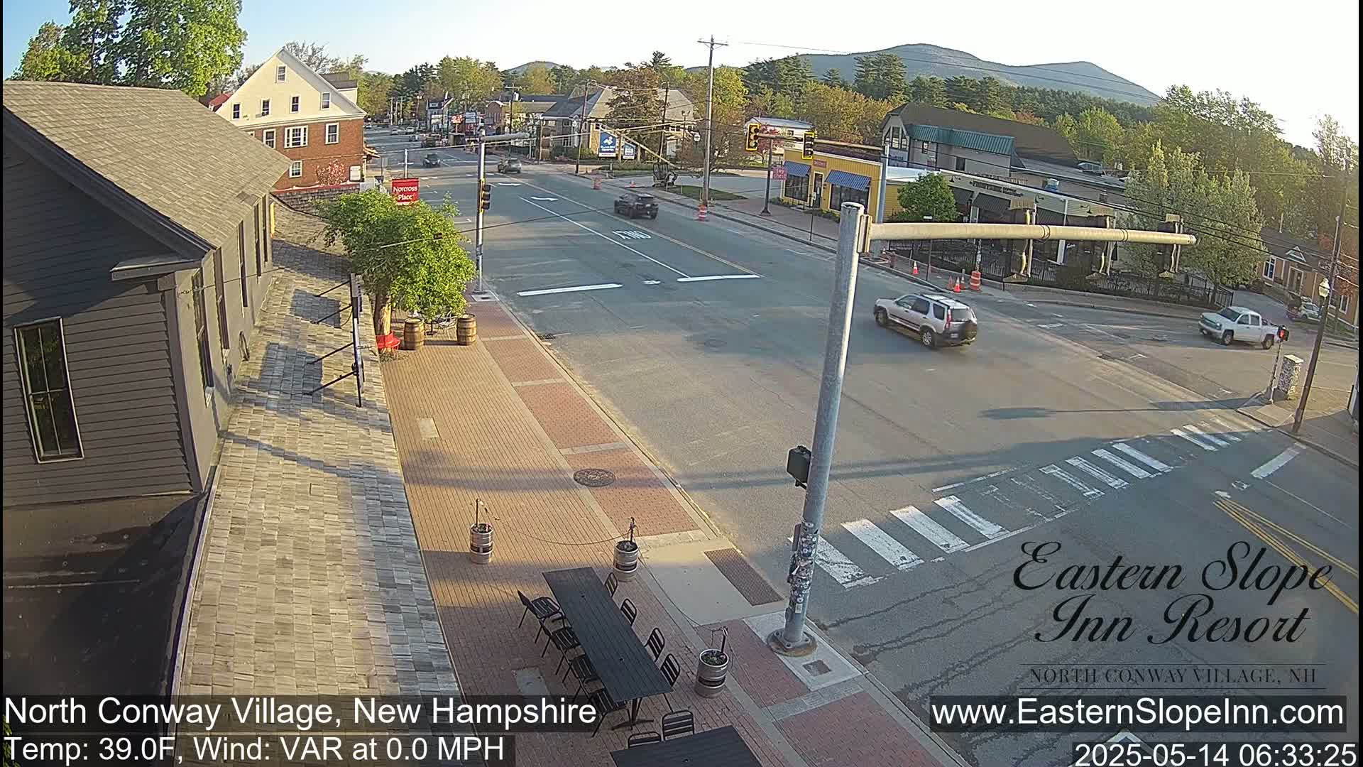 A sunny street scene shows a quiet intersection in a small town with a few cars and pedestrians, and outdoor seating at a cafe next to a brick building.