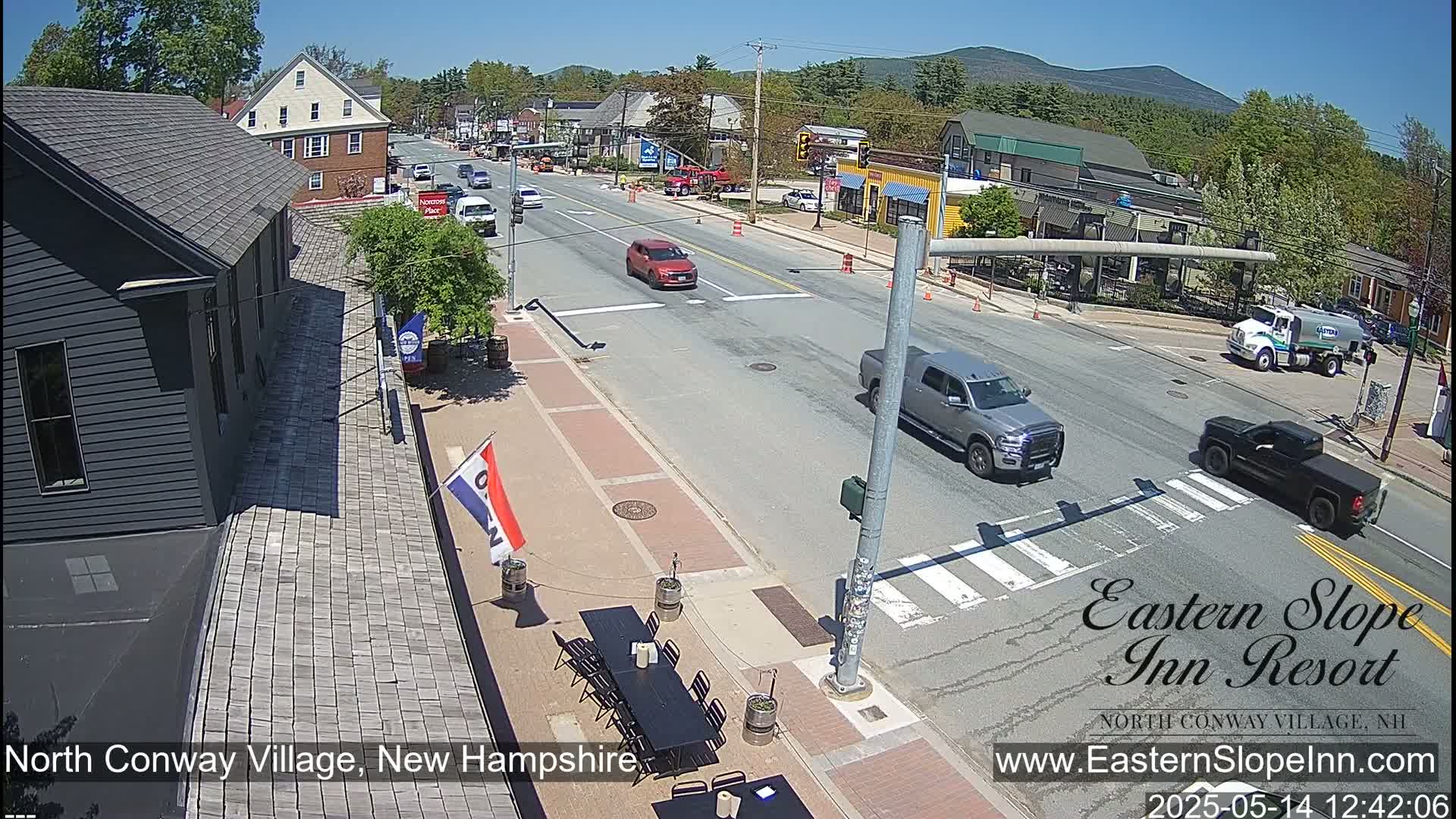 A sunny street scene in a small town shows several vehicles at an intersection, with outdoor seating at a restaurant visible on the side of the road.
