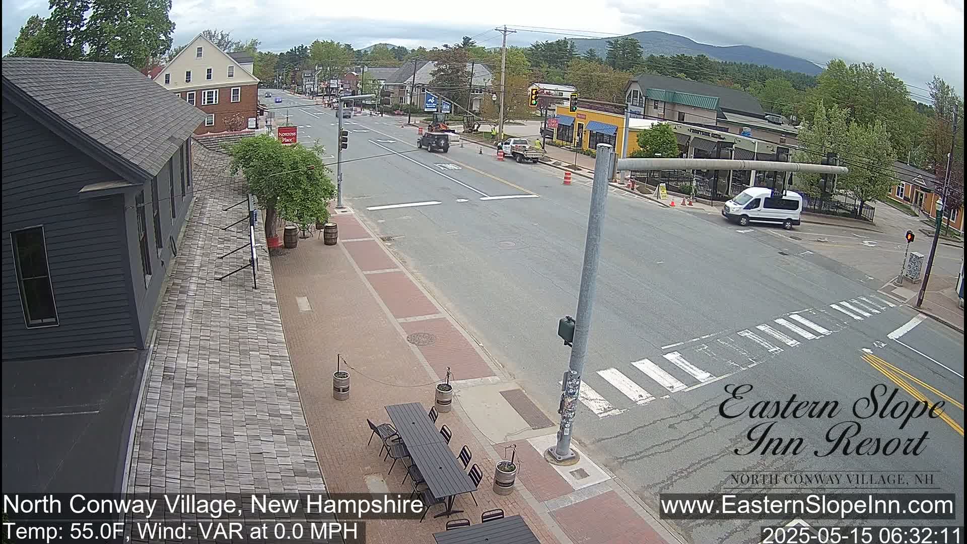 A mostly empty street scene on a partly cloudy day shows a village intersection with several buildings and some cars; outdoor seating is visible near a dark-colored building.