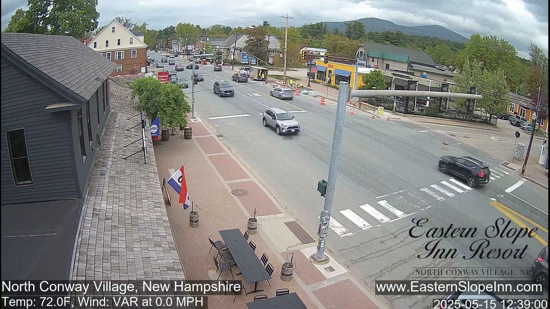 A partly cloudy day shows a street scene with several cars, a pedestrian crossing, and outdoor seating at a restaurant; mountains are visible in the background.