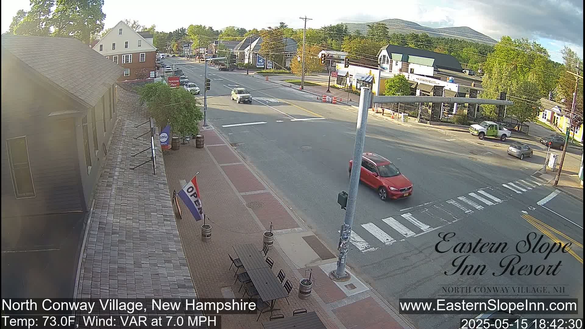 A partly cloudy day shows a street scene with several cars, a pedestrian crossing, and outdoor seating at a restaurant; mountains are visible in the background.