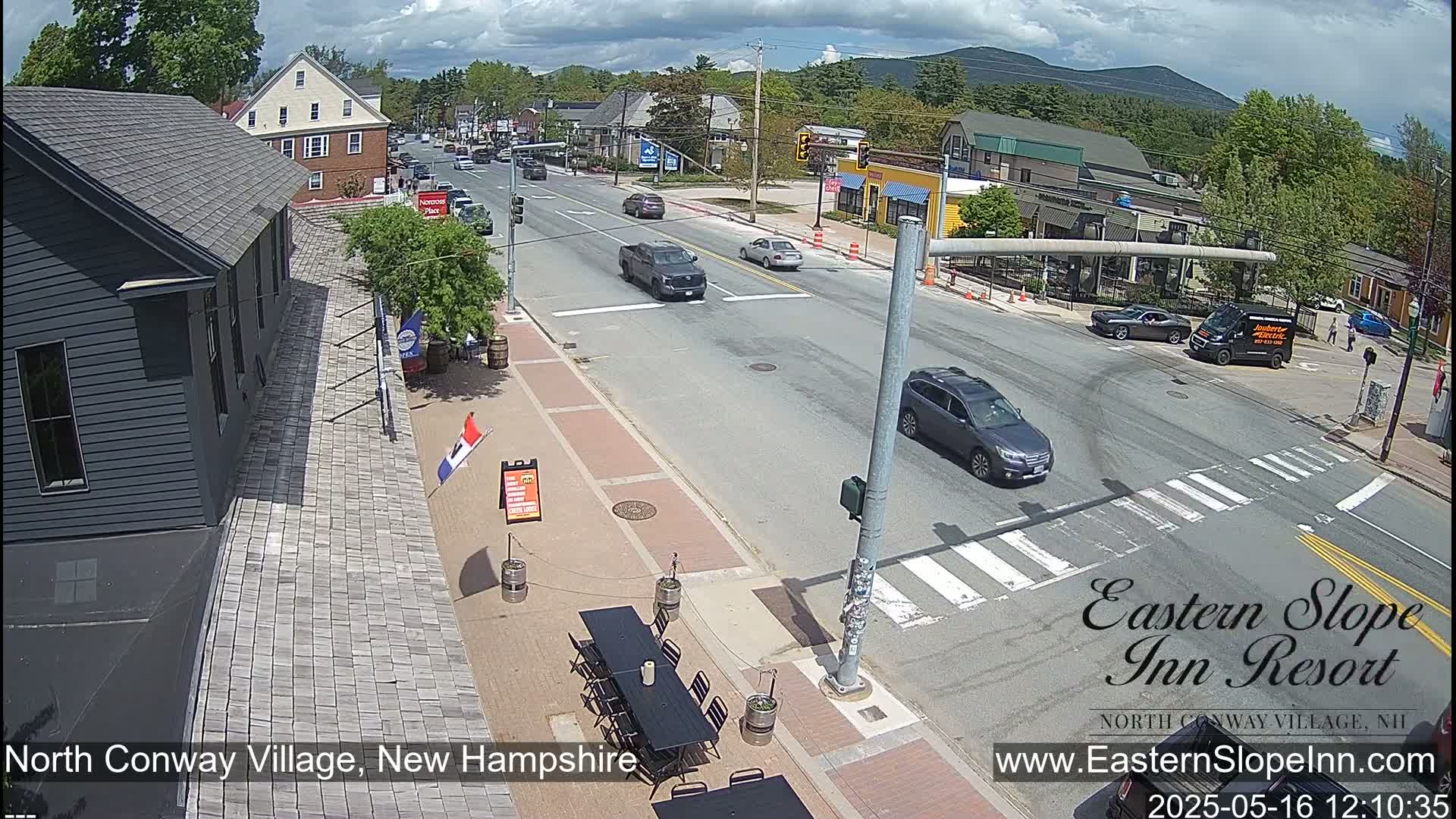 A partly cloudy day shows a street scene in a town with cars driving on a road that intersects with another, with buildings and outdoor seating visible on the sides.