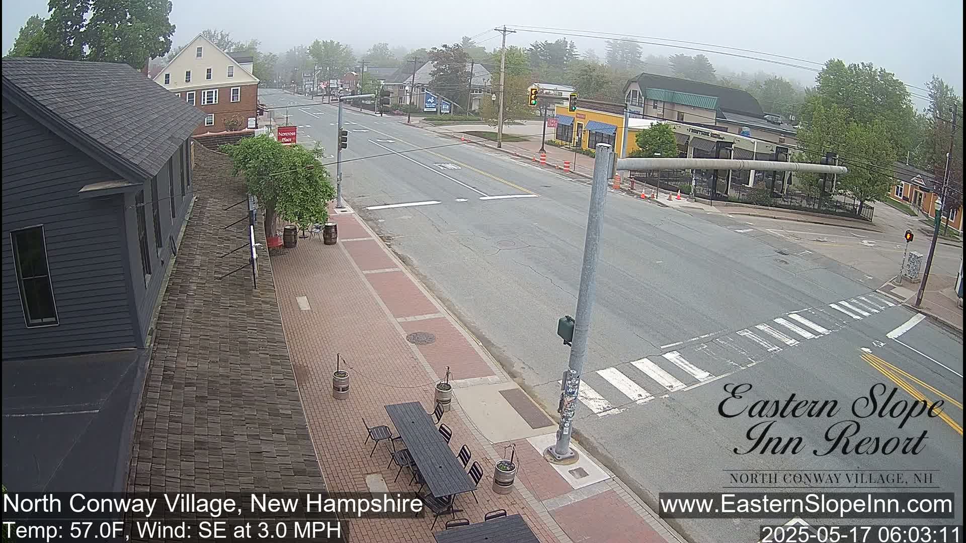 A mostly empty street scene on a foggy day shows a brick-paved sidewalk with outdoor seating in front of a dark-colored building, across from a road intersection with light traffic and several other buildings visible.