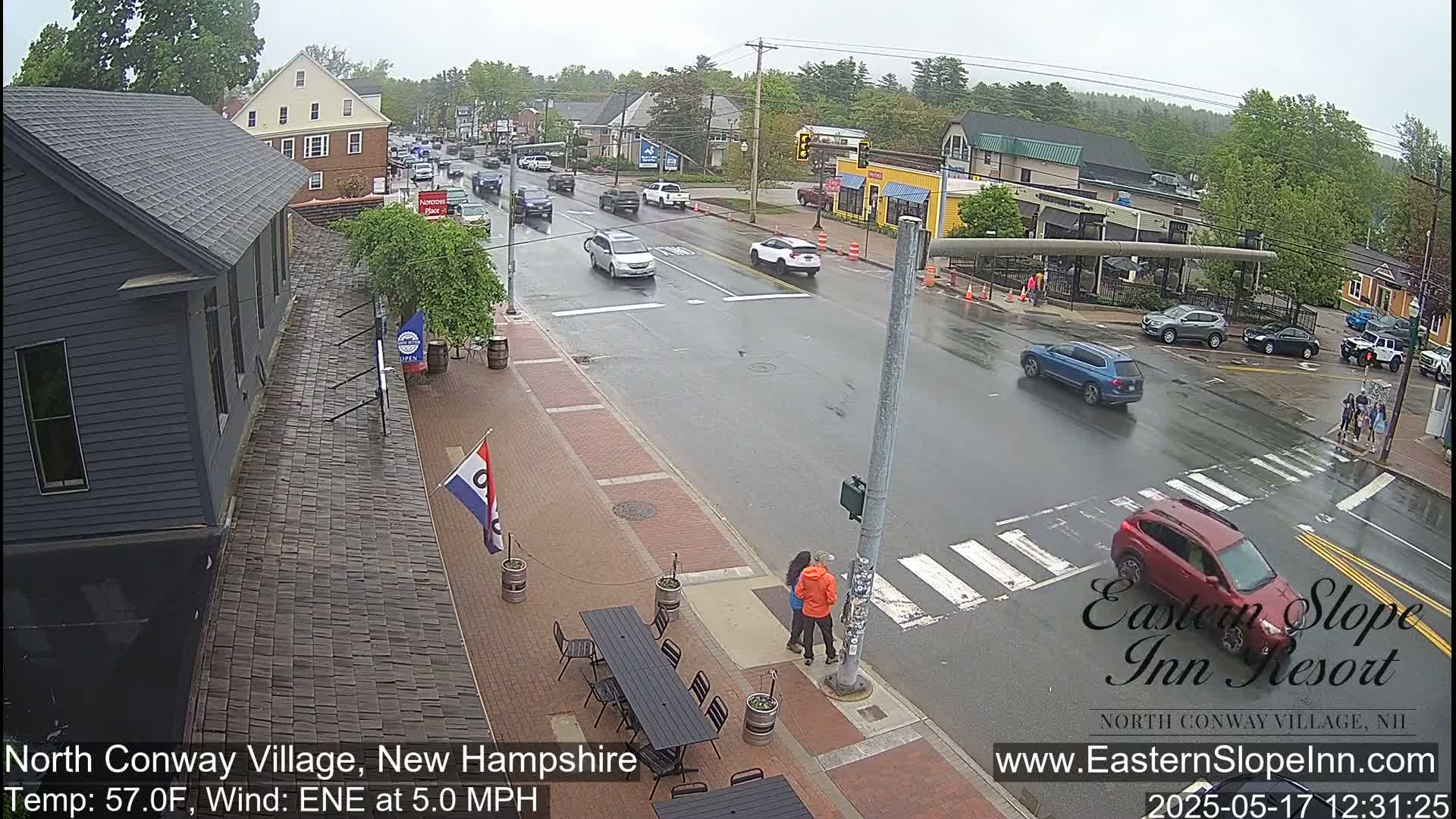 A rain-slicked street scene shows several cars at an intersection in a small town, with people standing on a sidewalk near outdoor tables in front of a dark-colored building.