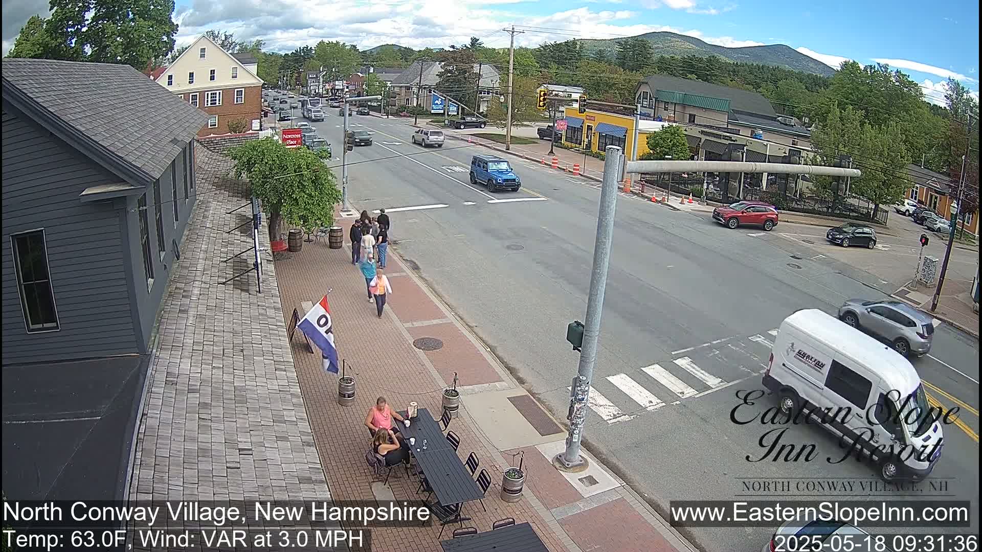 A street scene on a partly sunny day shows people walking and sitting at outdoor tables near buildings, with cars driving on a road in front of them and mountains in the distance.