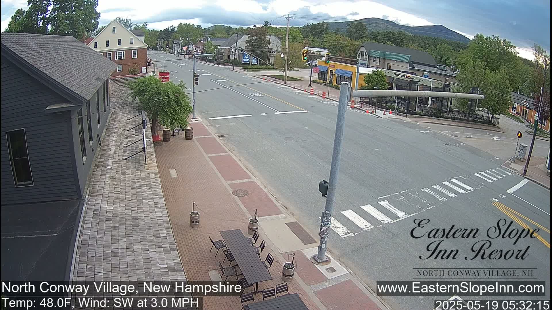 A mostly sunny street scene in a small town shows a road intersection with pedestrian crosswalks, buildings, and outdoor seating.