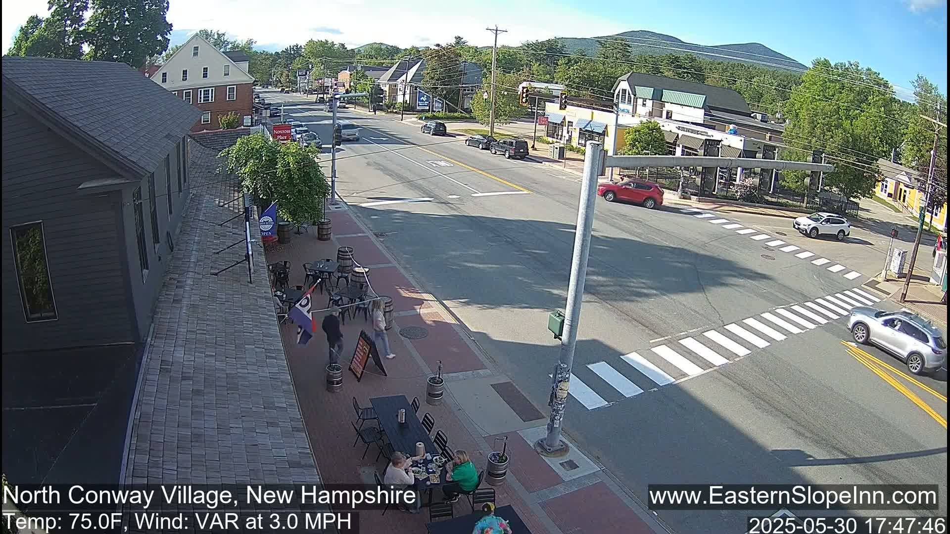 A sunny street scene shows a crossroads in a small town with several cars, pedestrians on a sidewalk patio, and buildings on either side of the road.