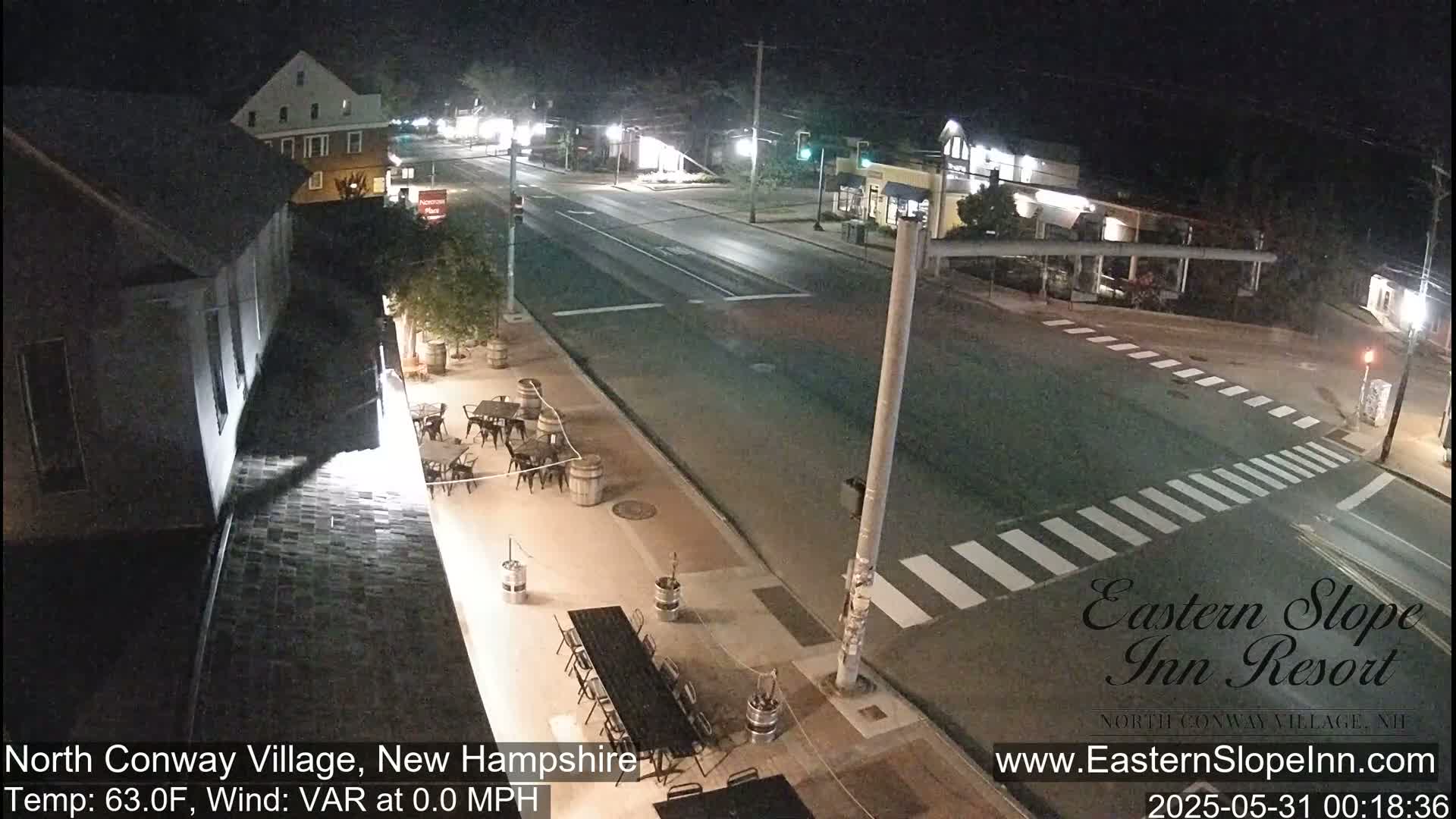 A nighttime street scene shows a mostly empty intersection with a pedestrian crosswalk, outdoor seating areas with tables and chairs, and several buildings along the road.