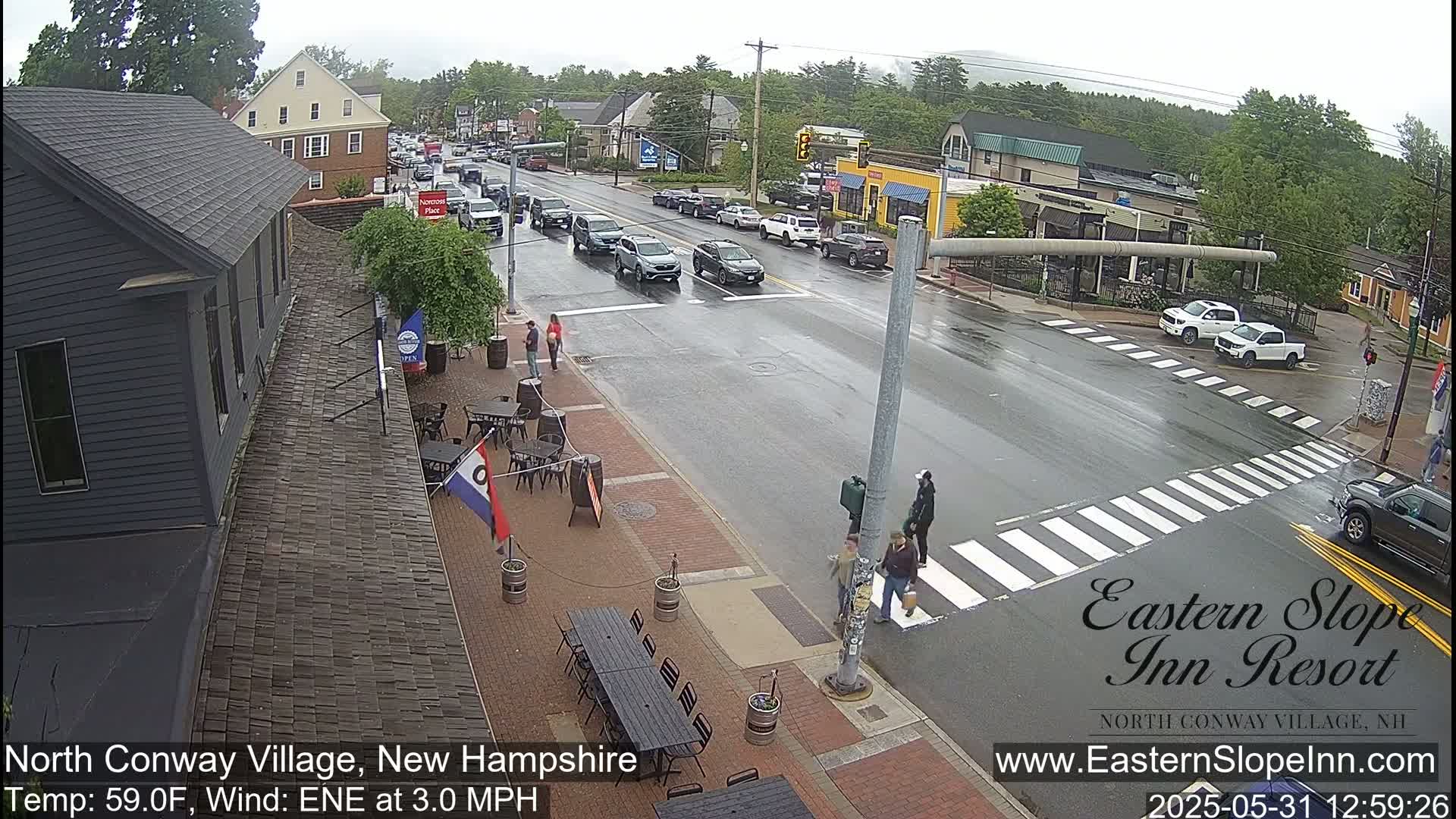 A wet street scene shows several cars driving past a crosswalk where pedestrians are walking, alongside buildings with outdoor seating areas.