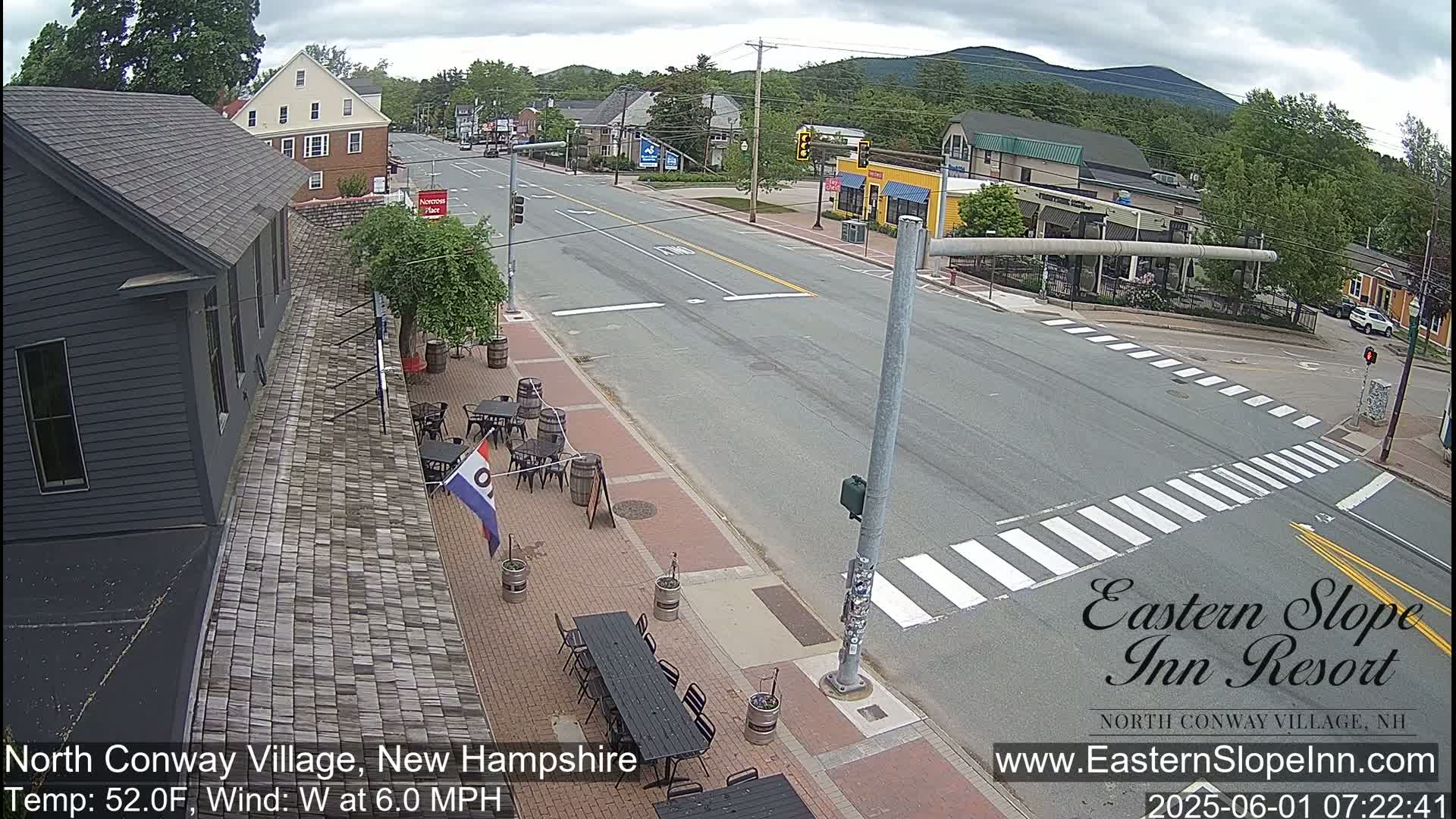 A mostly sunny view of a town's street with outdoor patio seating, light traffic, and trees under a partly cloudy sky.
