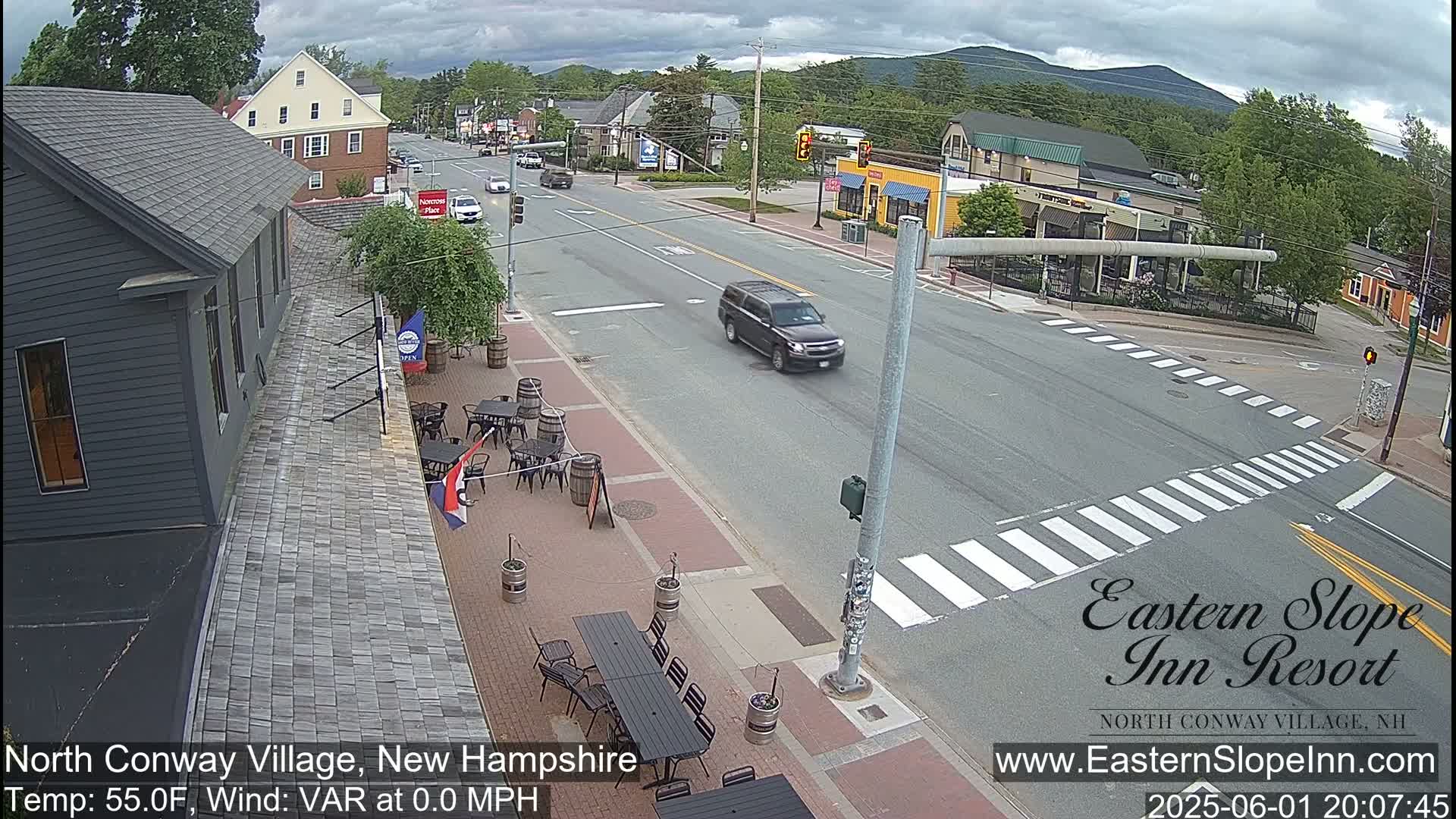 A mostly cloudy day shows a street scene with a dark-colored SUV driving on a road with crosswalks, alongside buildings and outdoor seating areas with several tables and chairs.