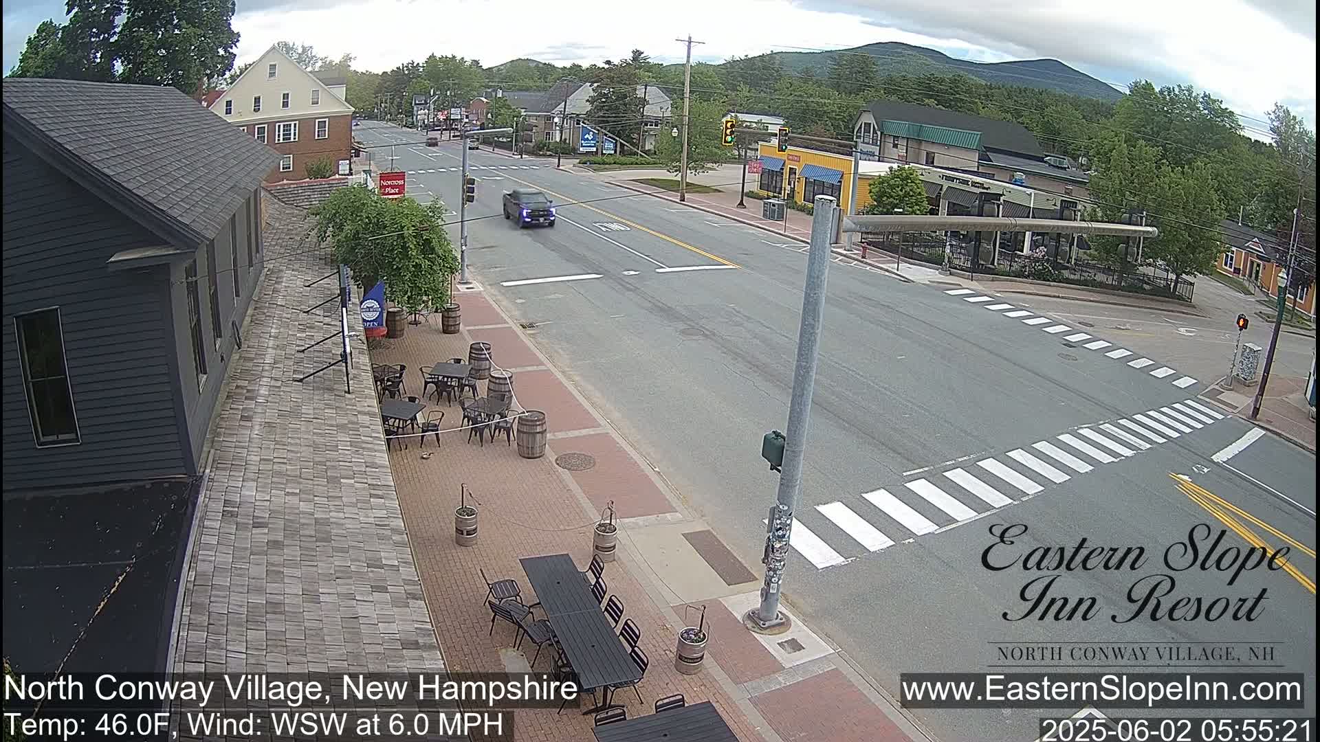 A mostly sunny street scene shows a paved road with crosswalks and a few cars, bordered by buildings, trees, and outdoor patio seating.