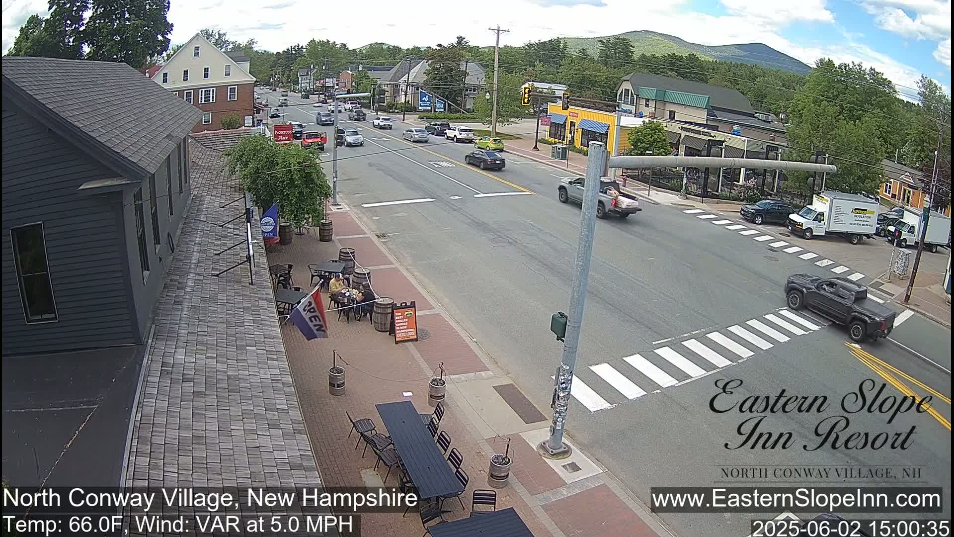 A street scene on a sunny day shows a town intersection with cars and pedestrians, alongside outdoor seating at a restaurant.