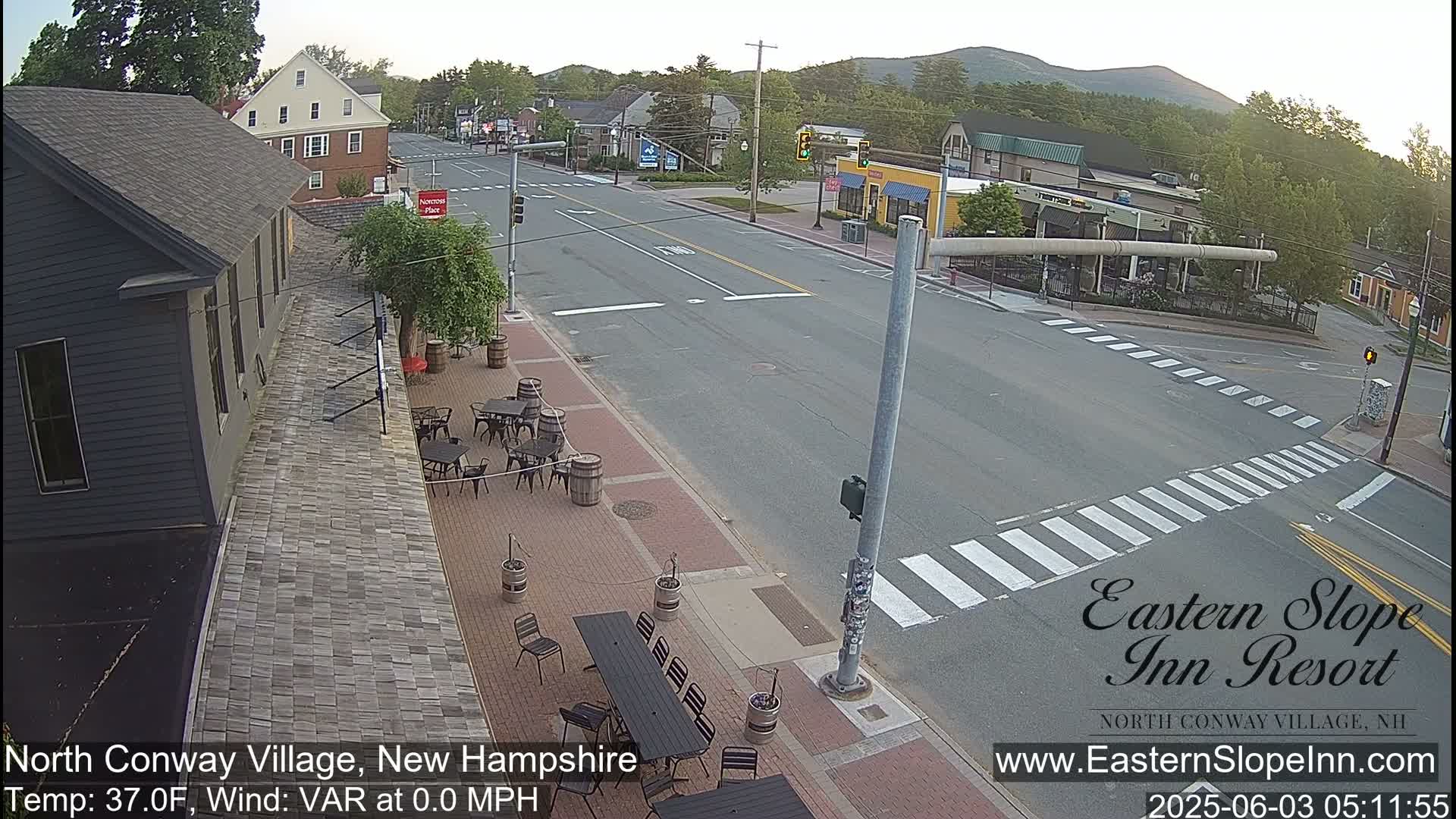 A street scene in a village on a partly sunny morning shows a quiet intersection with outdoor seating at a restaurant next to a building, and mountains in the background.