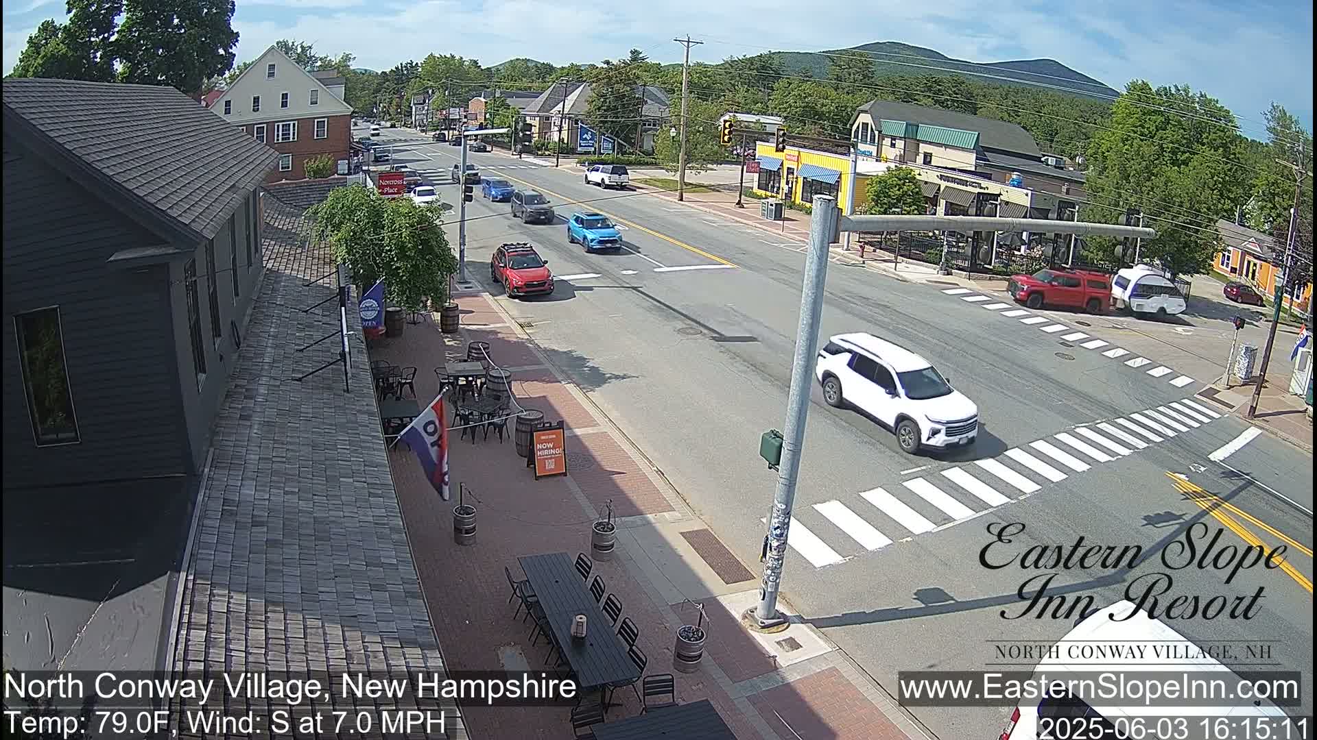 A sunny street scene shows several cars driving on a road that intersects with a crosswalk, and outdoor seating areas at a business with a partially visible sign.