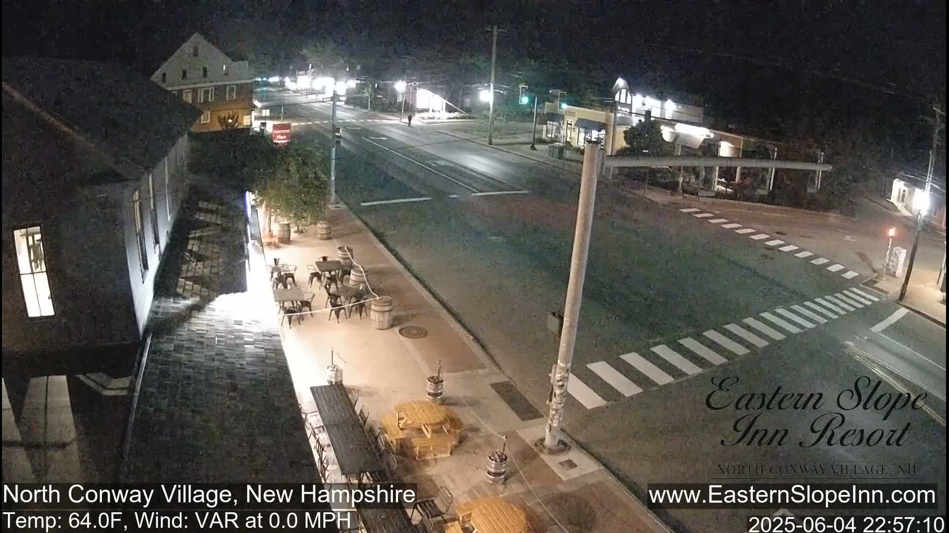 A nighttime street scene shows a mostly empty road intersection with outdoor seating at a restaurant, under calm, clear skies.