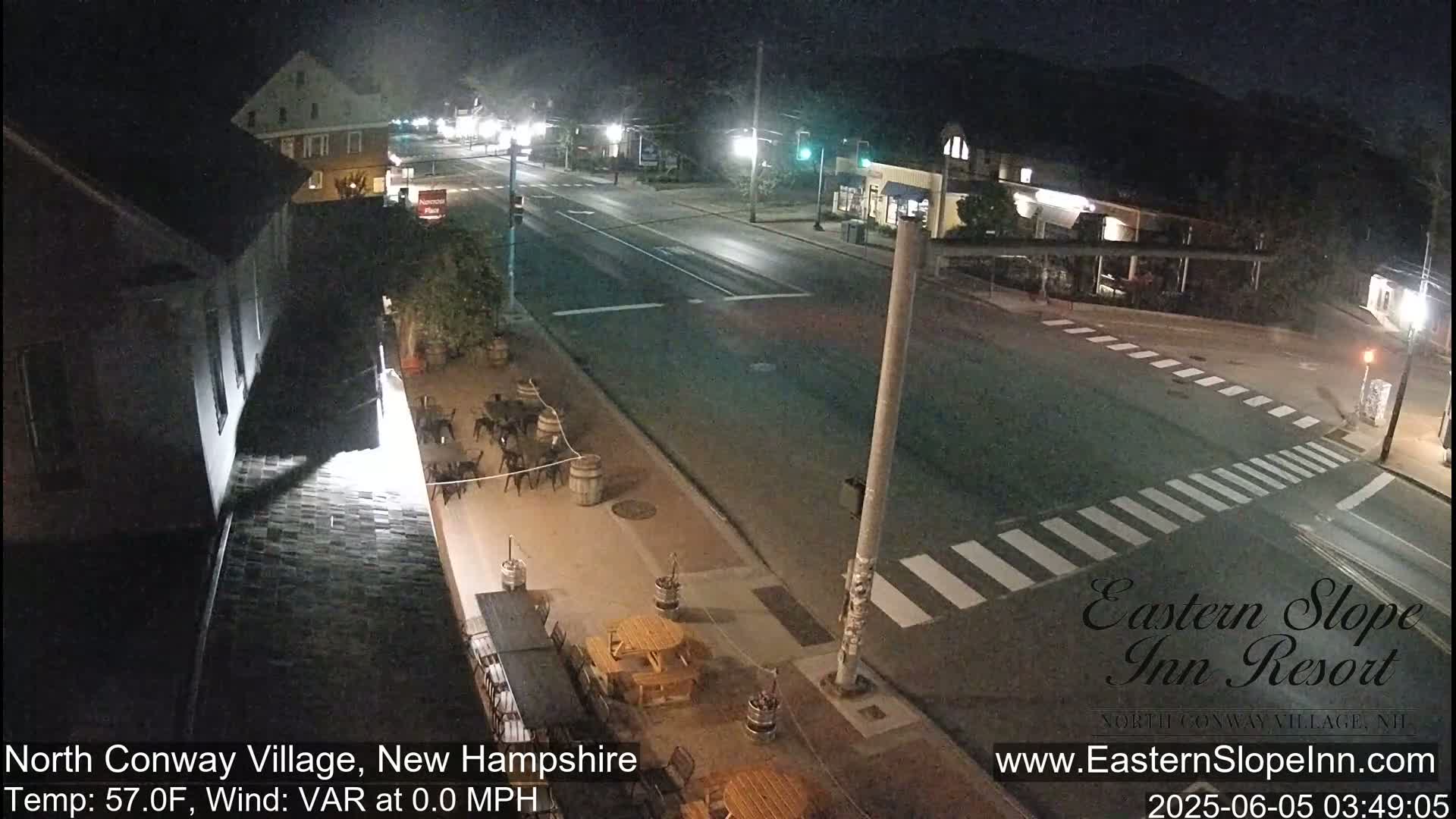 A nighttime street scene shows a crosswalk, a road with minimal traffic, and an outdoor patio area with tables and chairs.