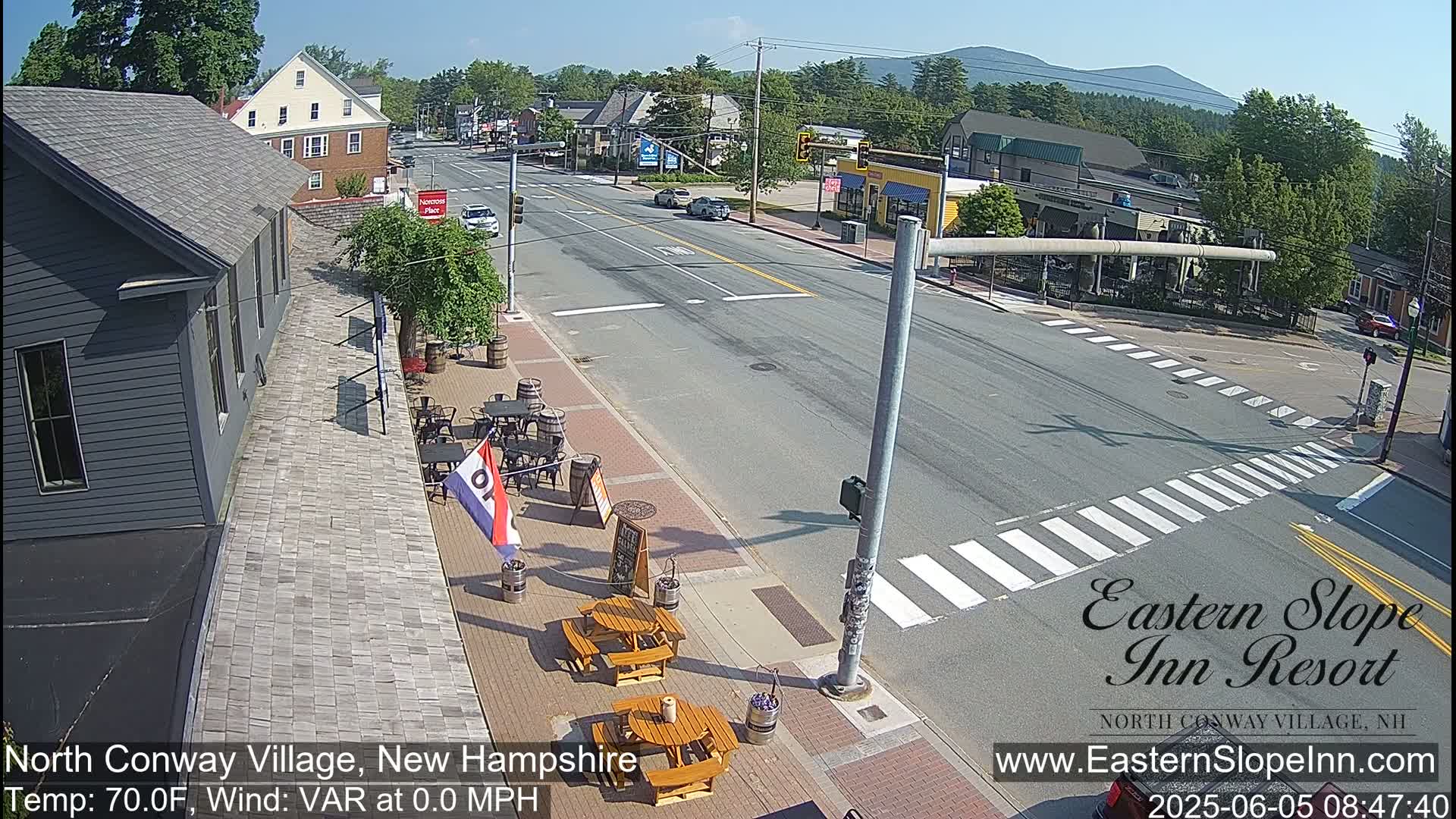 A sunny view of a town street intersection with a few cars, outdoor seating at a restaurant, and mountains in the distance.