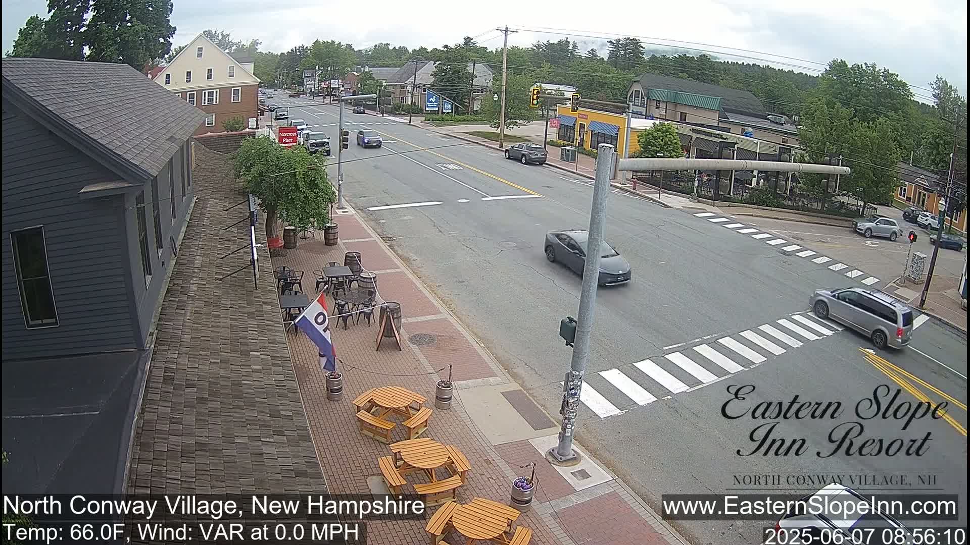 A street scene shows a mostly empty road with several cars, a pedestrian crosswalk, outdoor seating at a restaurant, and several buildings under a partly cloudy sky.