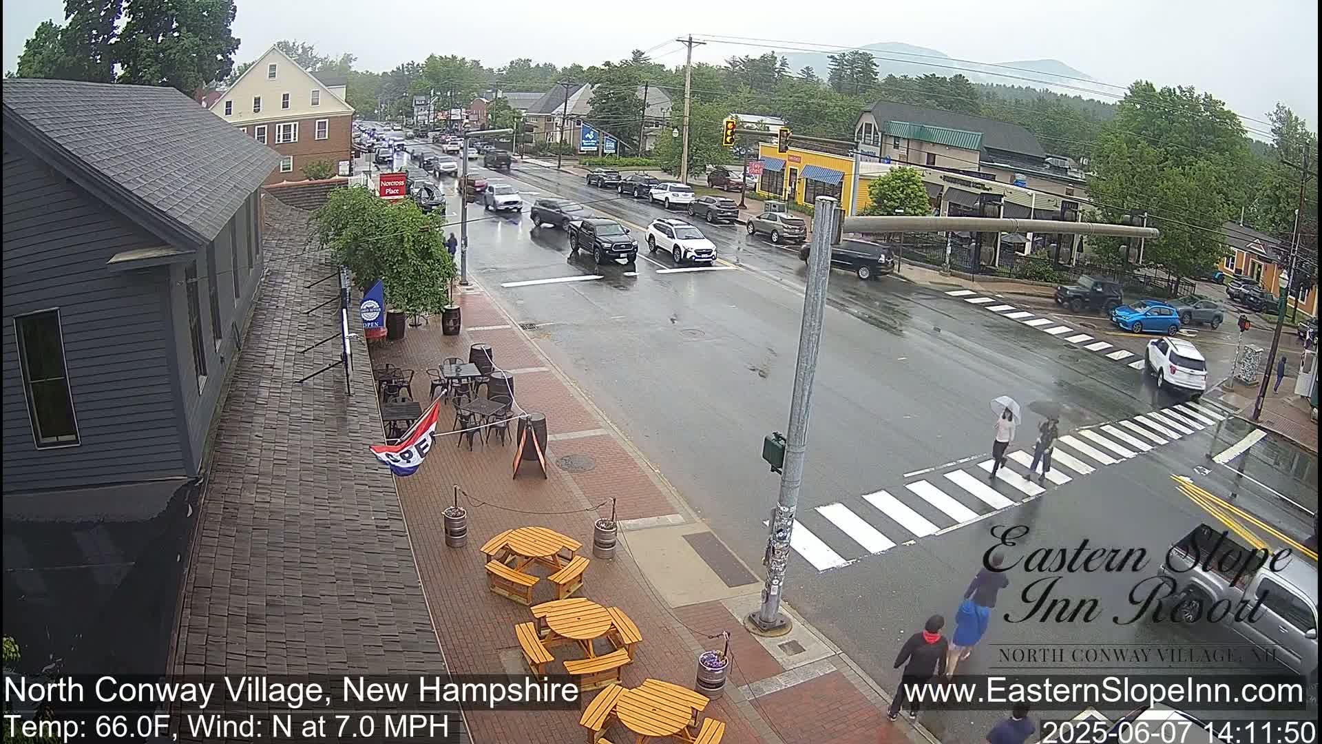 A rain-slicked street scene shows several vehicles driving through a town intersection, with pedestrians crossing a crosswalk and outdoor seating visible at a restaurant.