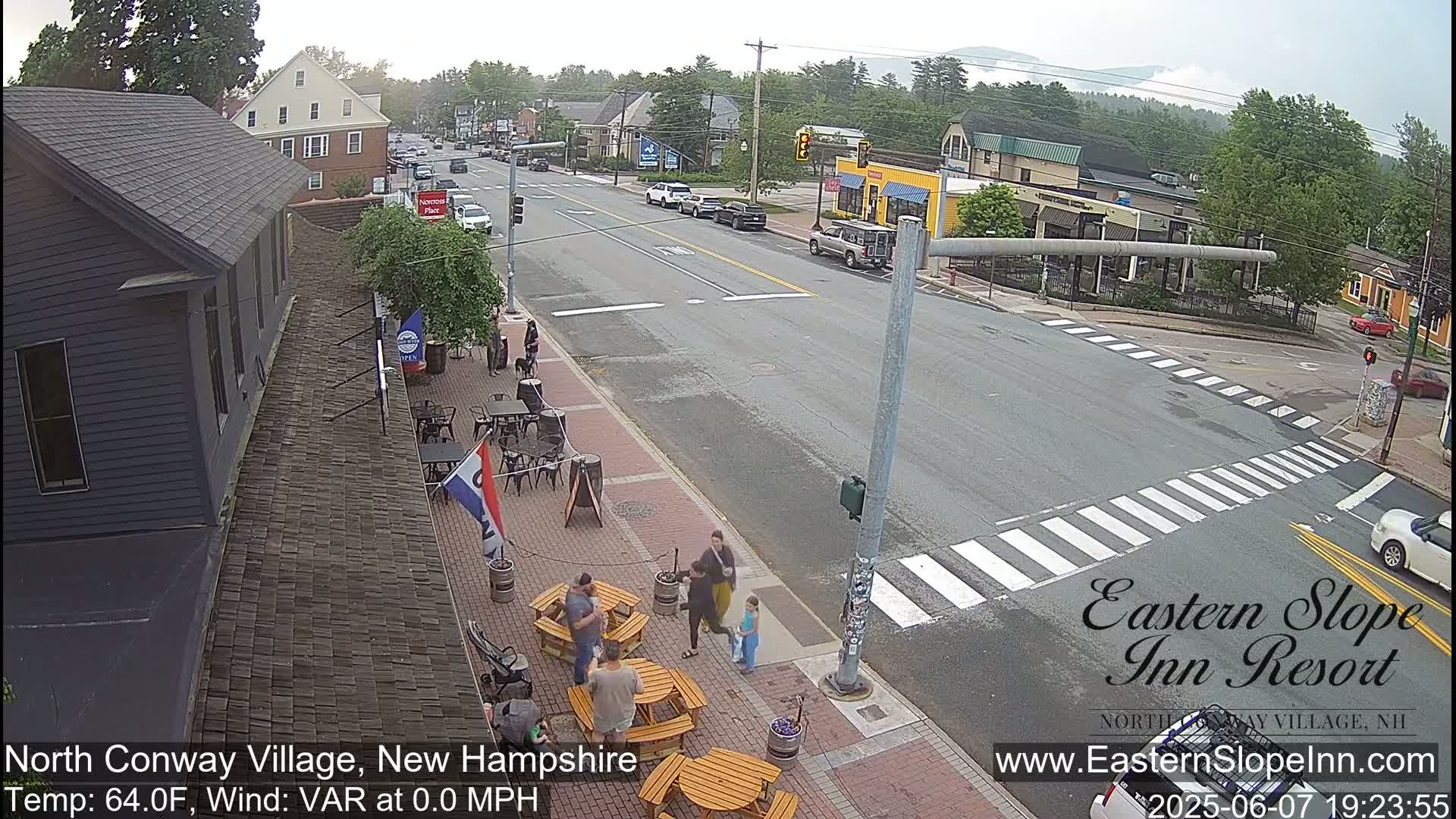 A street scene shows a partly sunny day with several people near outdoor seating at a restaurant, cars driving down a road, and buildings lining the street.