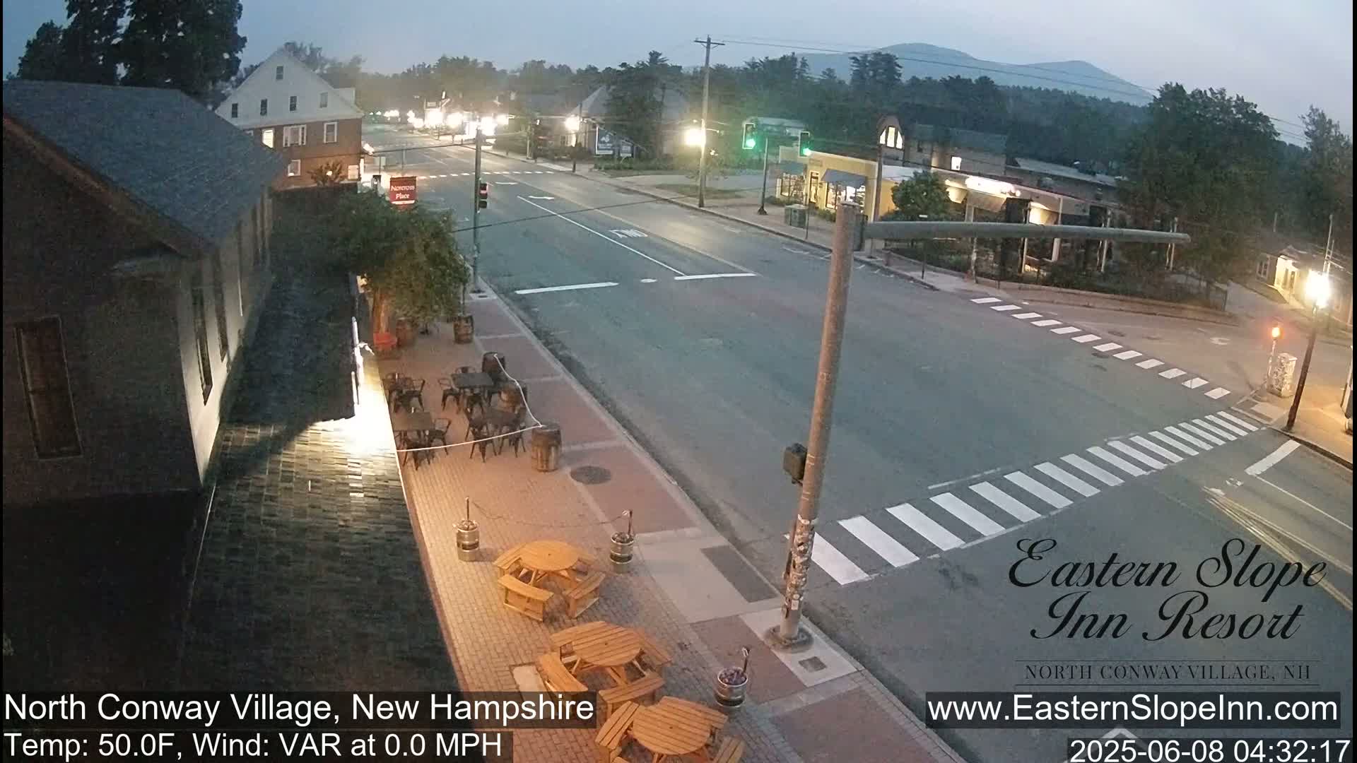 A mostly empty street scene at dawn shows a town with buildings, outdoor seating areas, and a crosswalk, under a clear sky.
