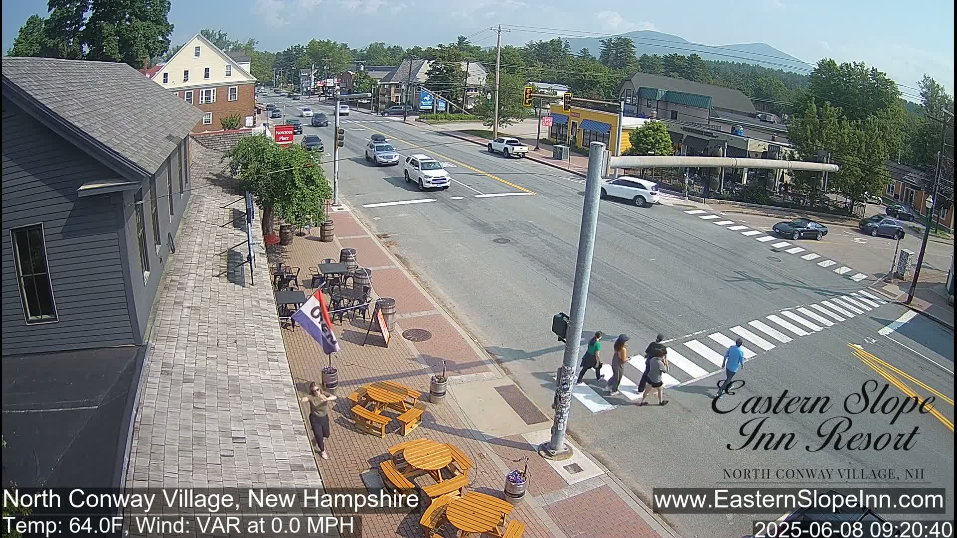 A sunny day shows a street scene with cars driving and pedestrians crossing a crosswalk, outdoor seating areas in front of buildings, and mountains in the background.