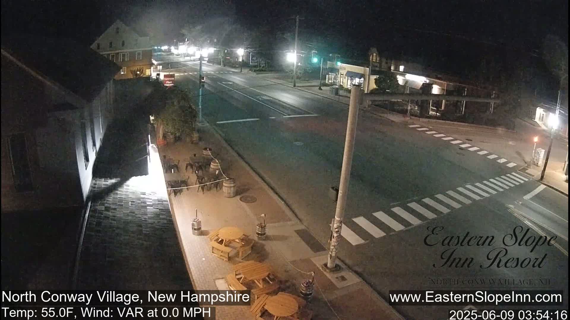 A nighttime, high-angle view of a mostly empty street with a crosswalk and outdoor seating area in front of a building.