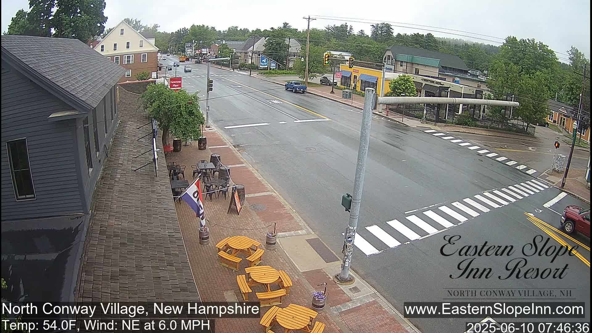 A wet street scene shows a road intersection with several buildings, outdoor seating at a restaurant, and a few cars.