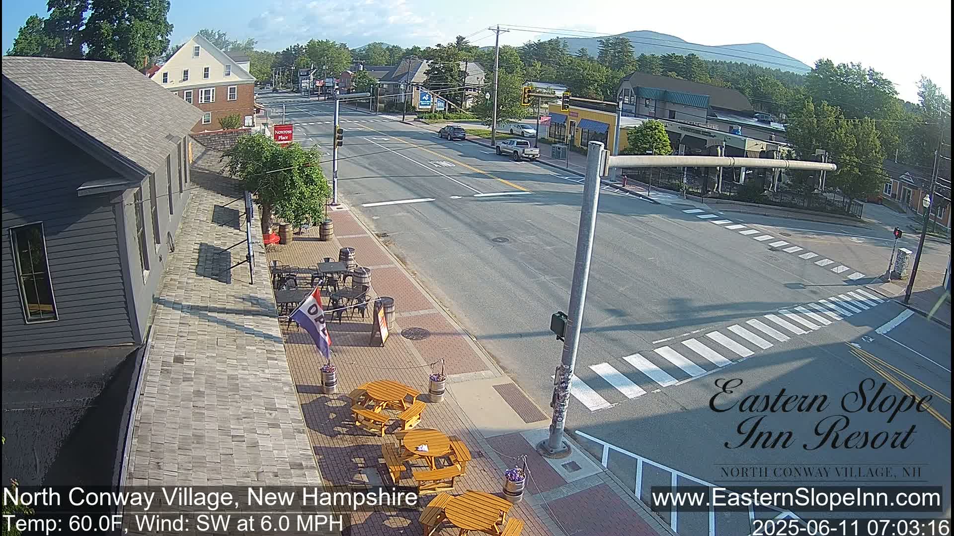 A sunny street scene in a town shows a street intersection with a few cars, pedestrian crosswalks, and outdoor seating at a restaurant next to a gray building, with mountains visible in the background.