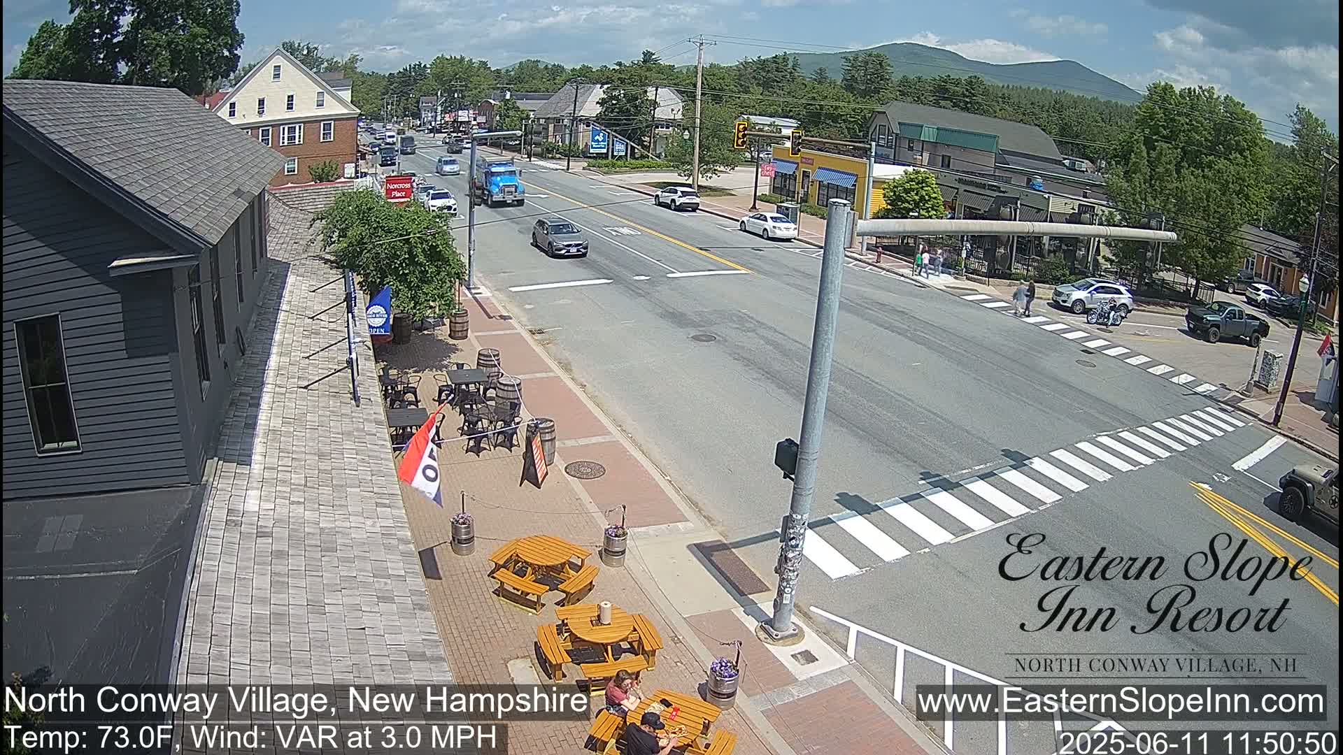 A sunny street scene in a village shows cars driving on a road that intersects with a crosswalk where pedestrians are walking, with outdoor seating at a restaurant along the road, and mountains visible in the distance.