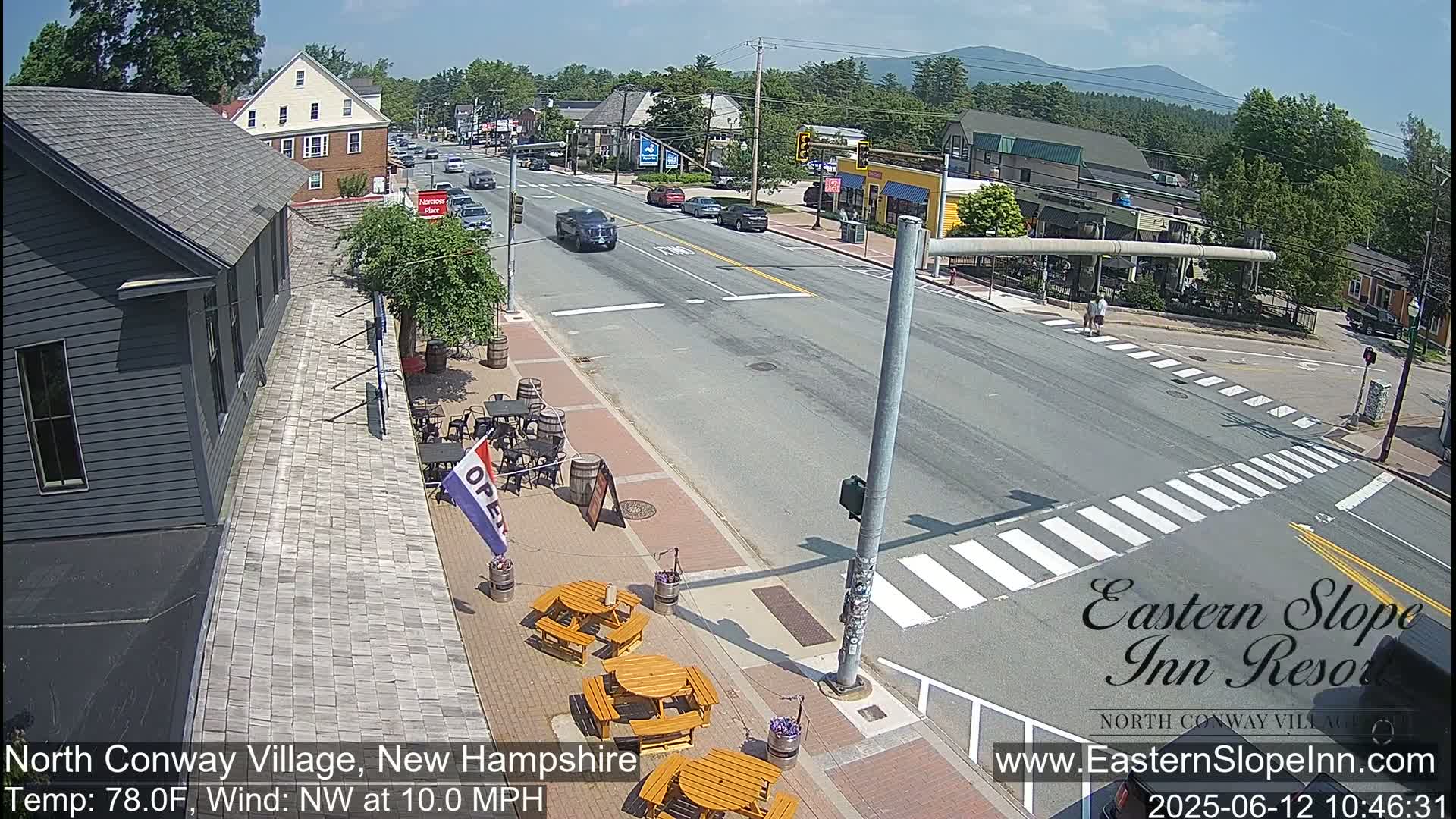 A sunny day shows a street scene in a small town with cars driving on a main road, pedestrians crossing at a crosswalk, and outdoor seating at a restaurant along the sidewalk.