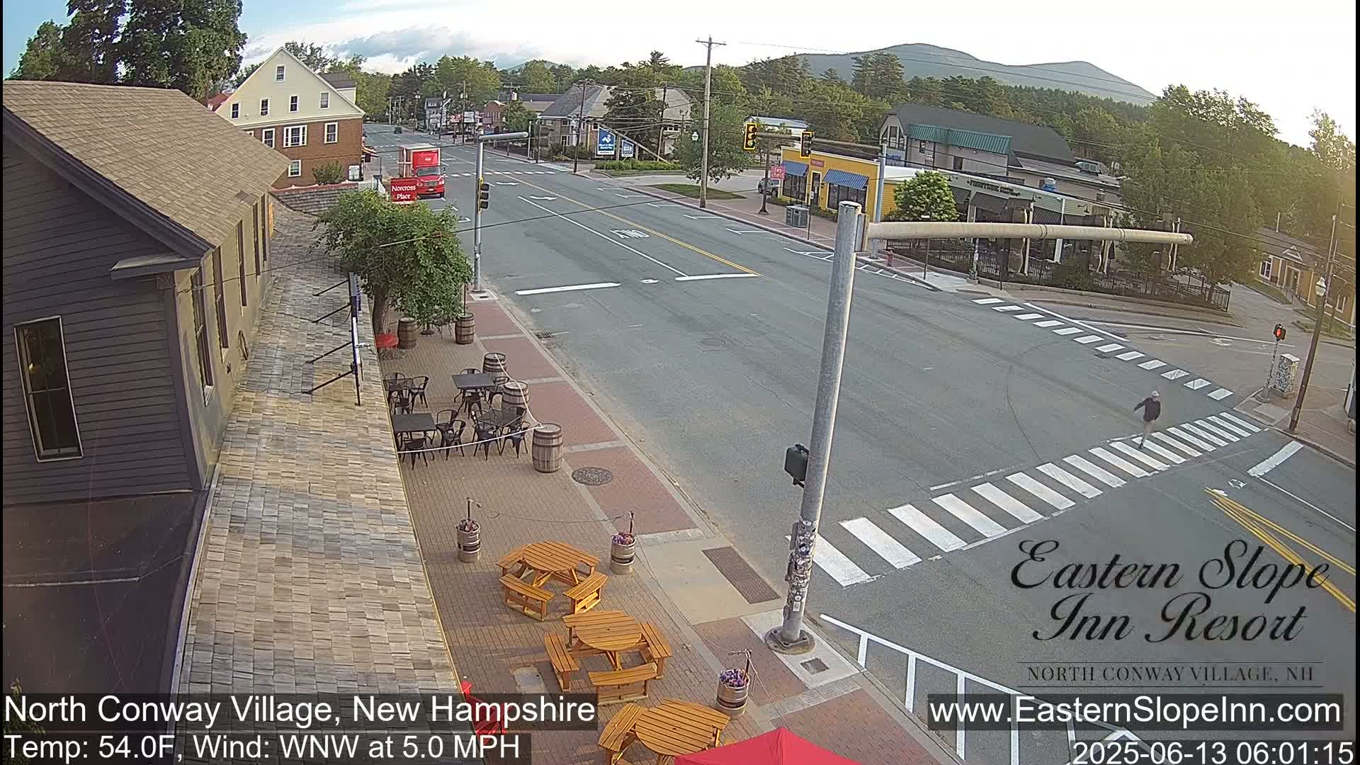 A street scene in a village shows a roadway intersection with outdoor seating at a restaurant, a pedestrian crossing, and a partly cloudy sky.