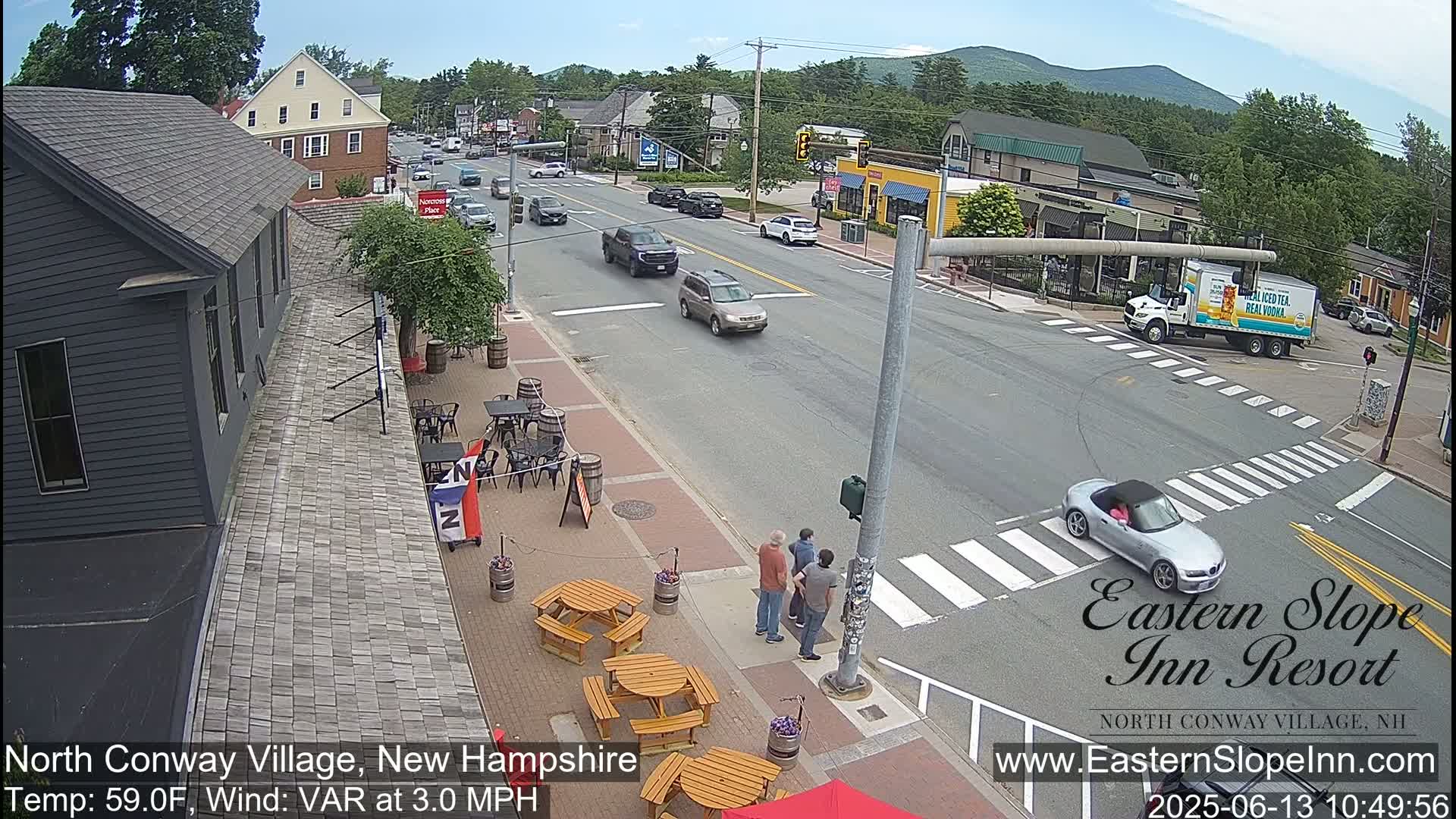 A sunny street scene shows several cars driving on a road lined with buildings, and pedestrians and outdoor seating areas are visible next to a building.