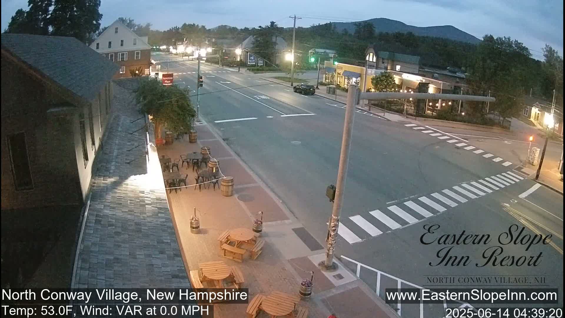 A mostly empty street scene at twilight shows a crossroads in a small town with outdoor seating at a restaurant and buildings lining the street under a clear sky.