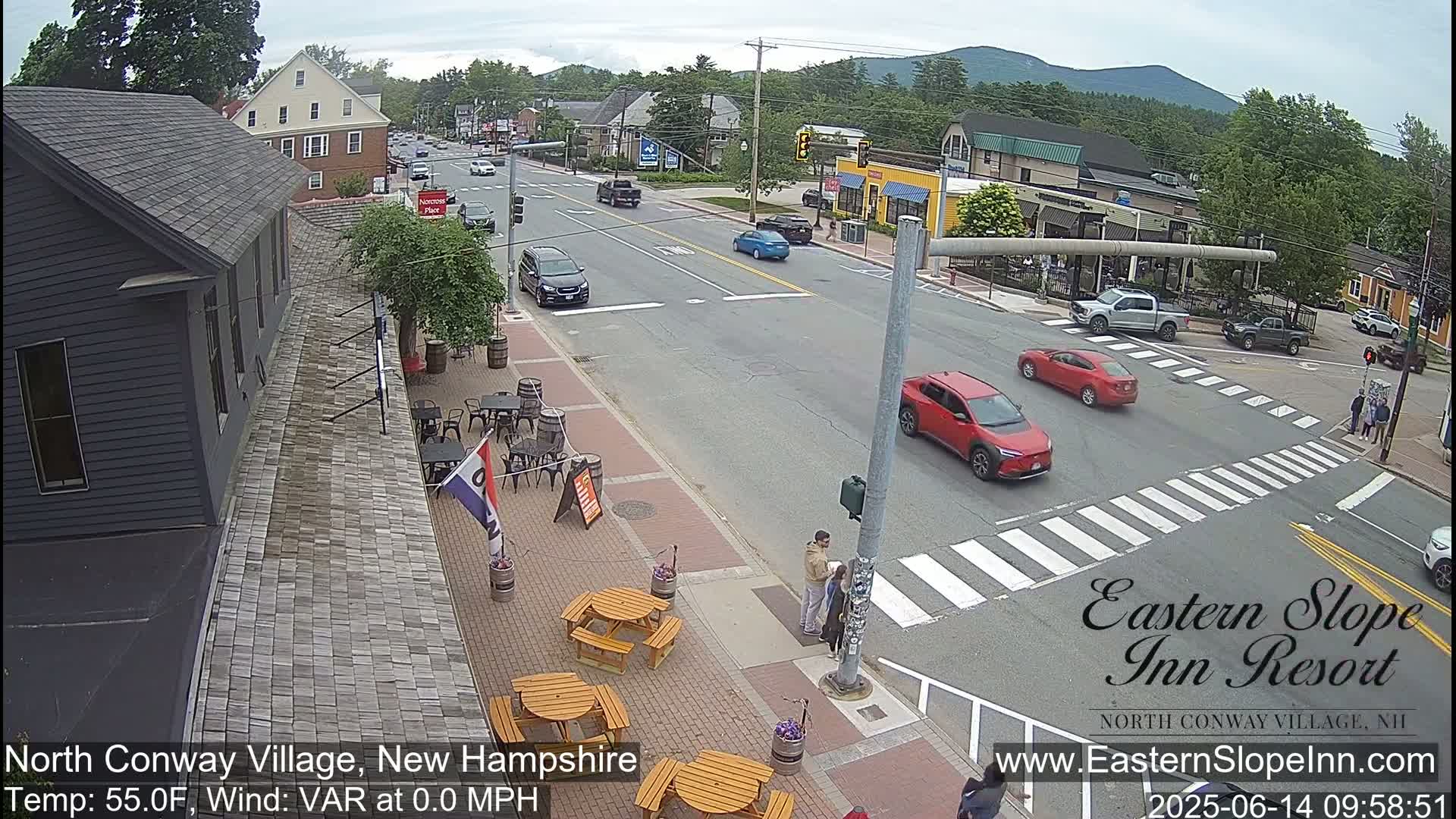 A partly cloudy day shows a street scene with cars at an intersection in a town with outdoor seating at a restaurant, and people near a crosswalk.