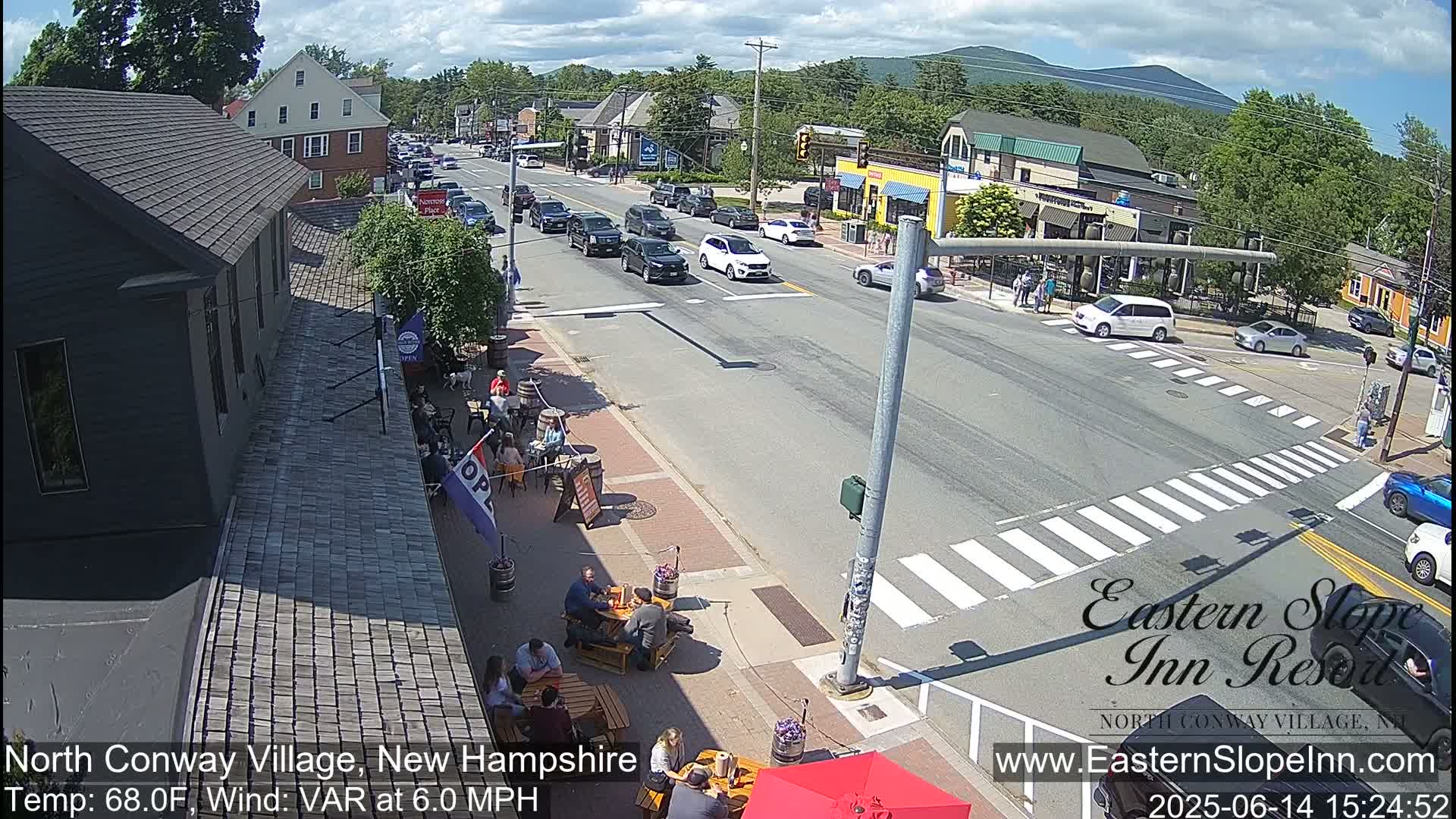 A partly cloudy day shows a street scene with cars at an intersection in a town with outdoor seating at a restaurant, and people near a crosswalk.