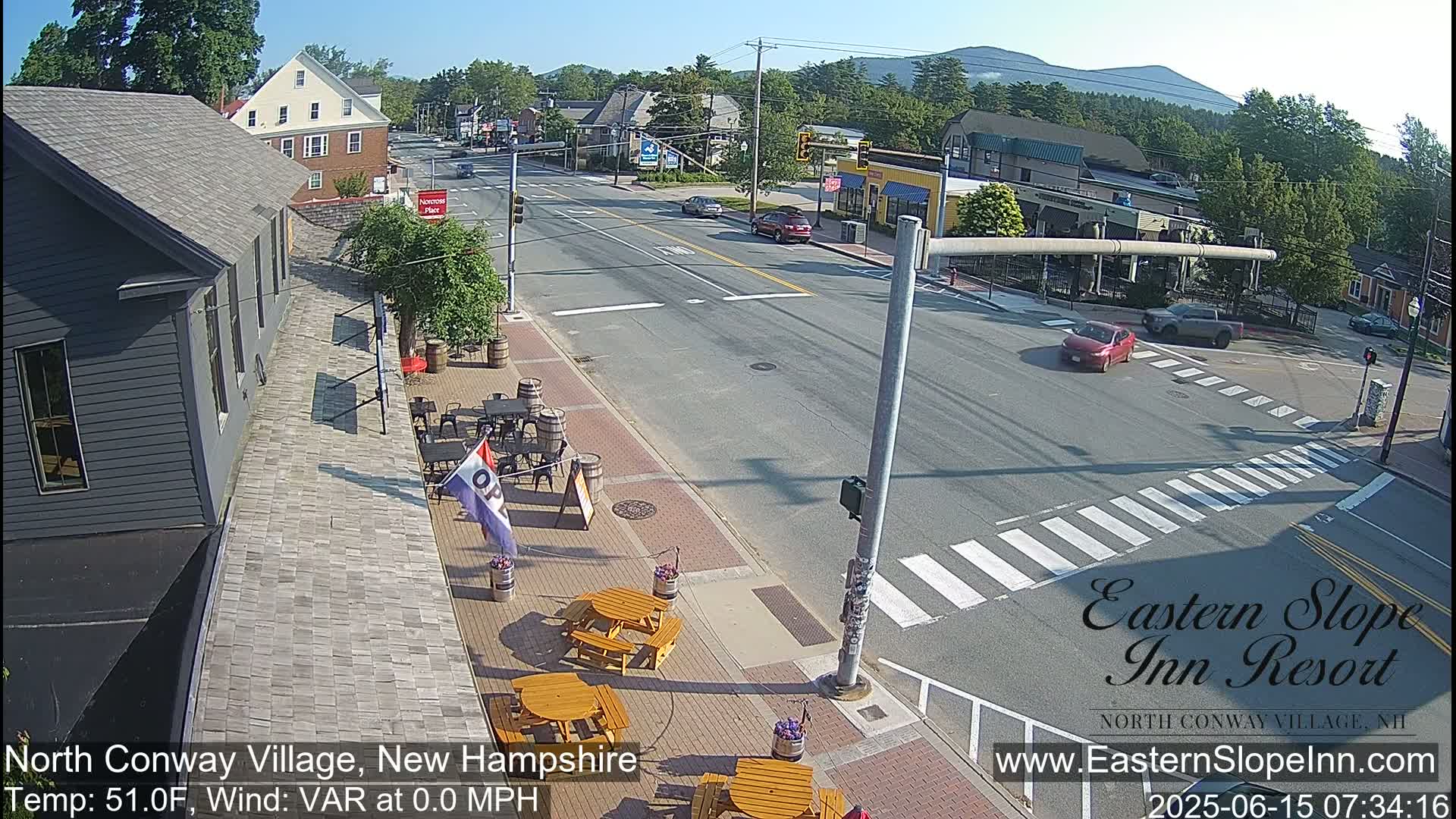 A sunny street scene shows a town intersection with cars and outdoor seating at a restaurant, under clear skies.