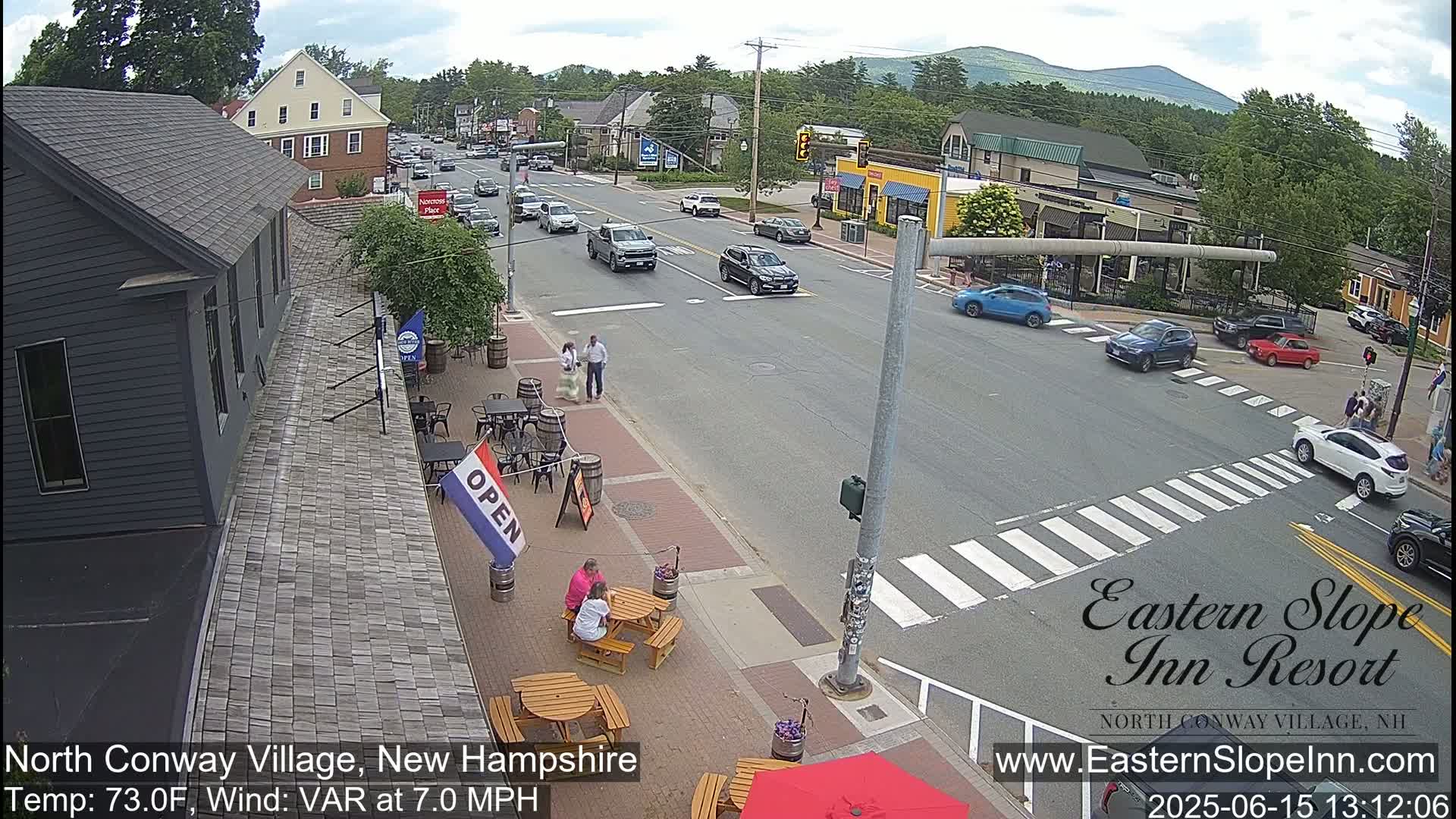 A street scene on a sunny day shows a town with cars driving on a road that intersects with a crosswalk, people walking and sitting at outdoor tables of a restaurant with a sign saying "open," and buildings along the road with mountains in the background.