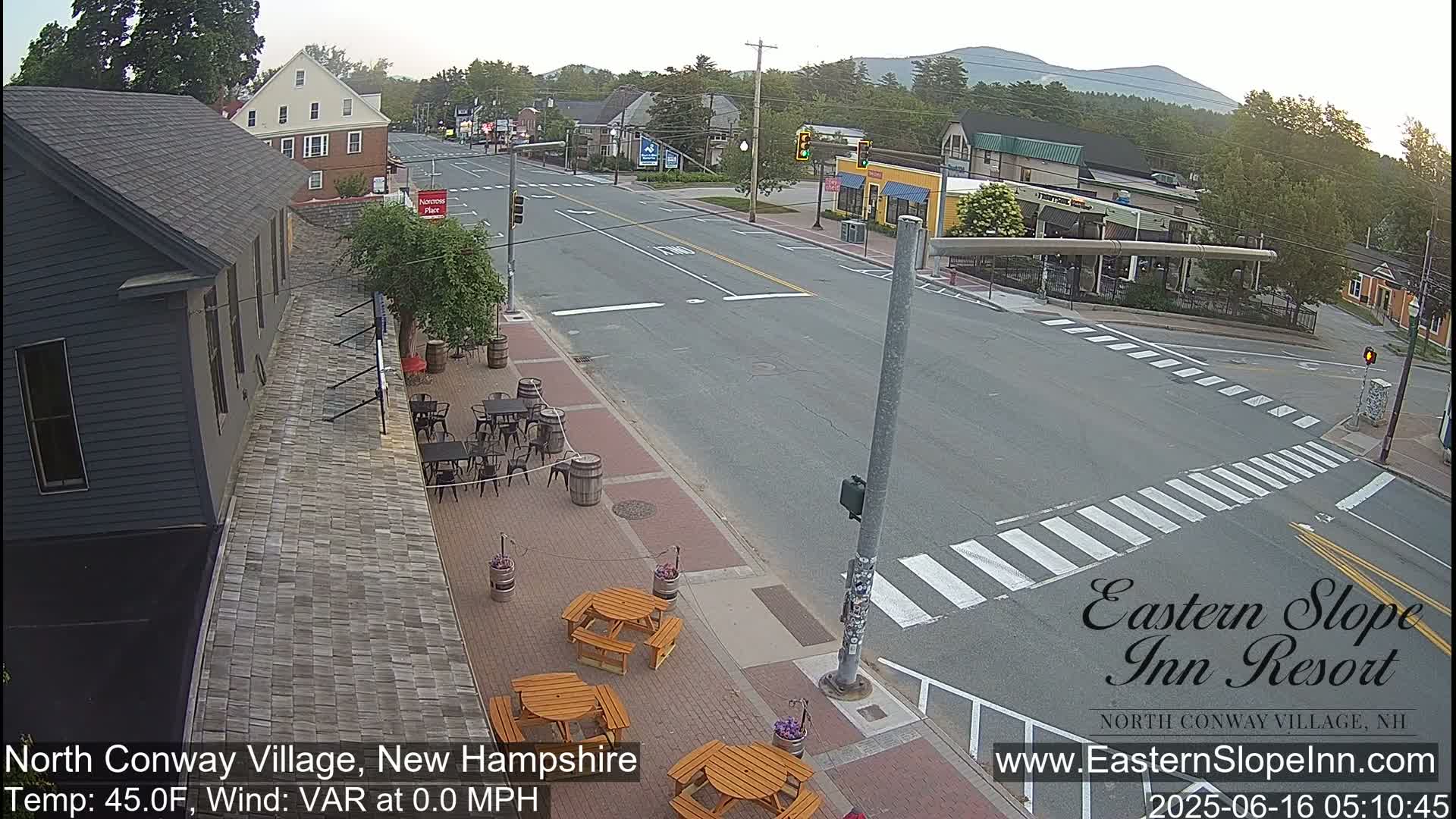 A mostly empty street scene in a village, showing a few buildings, outdoor seating areas, and a crosswalk, under partly sunny skies.
