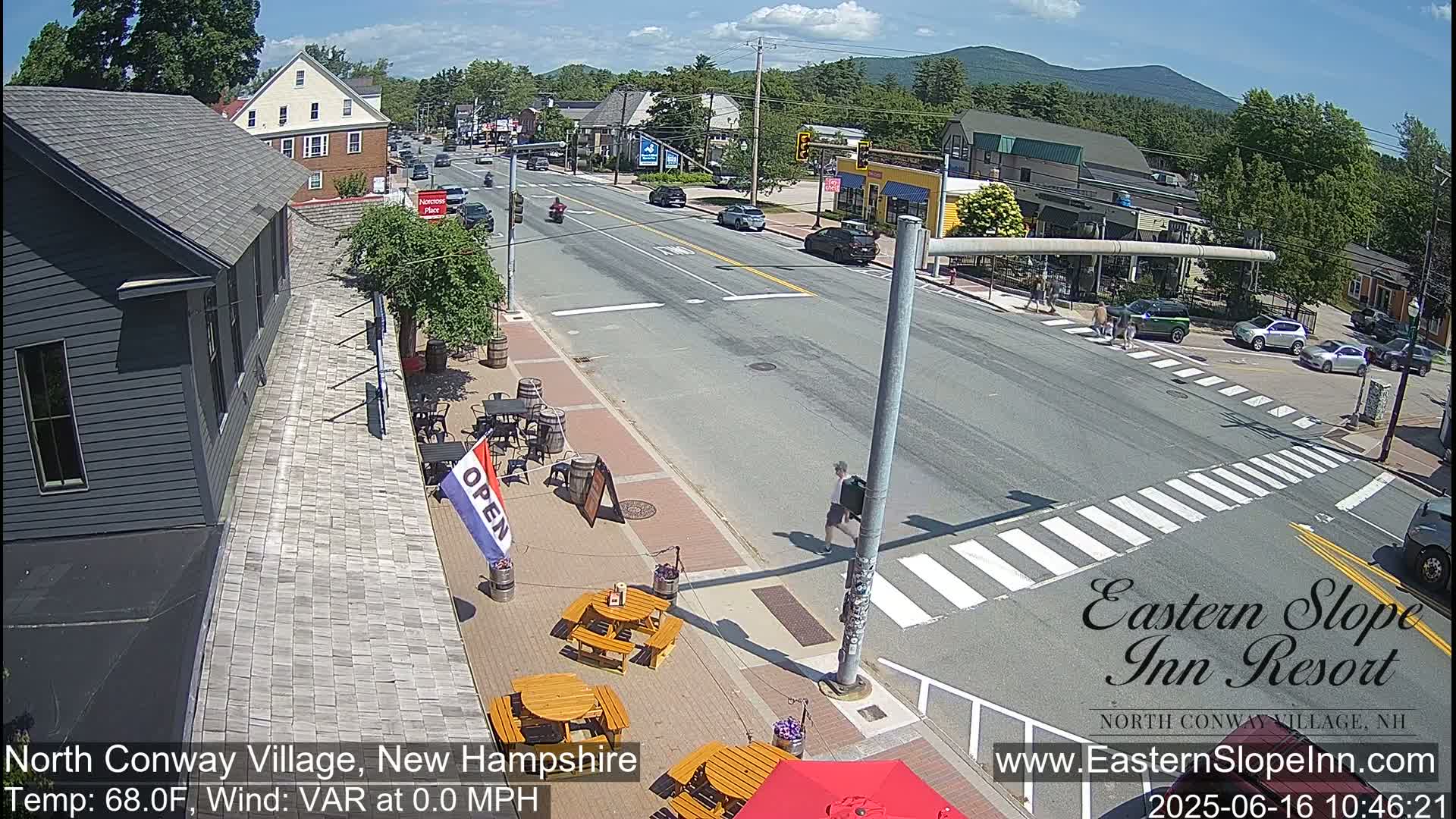 A sunny street scene shows a town's main road with cars and pedestrians, outdoor seating at a restaurant, and buildings lining the street, with mountains visible in the distance.