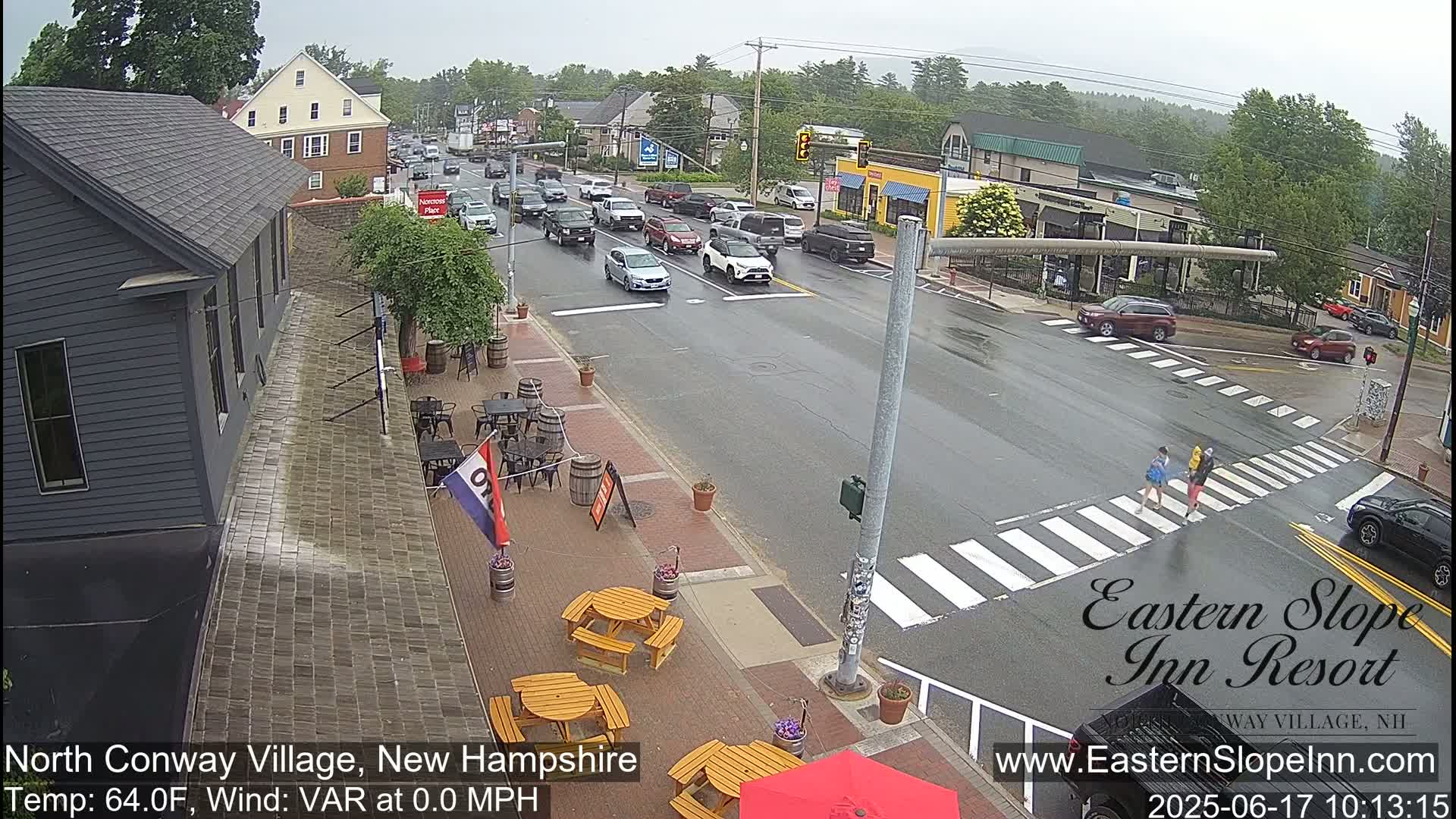 A wet street scene shows several cars driving through a town intersection on a rainy day, with outdoor seating at a restaurant visible to the left.