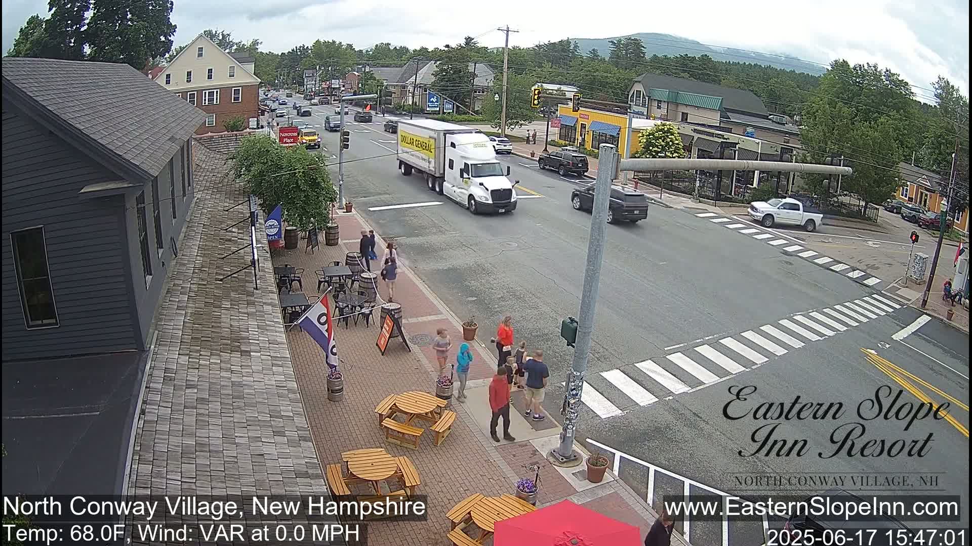 A street scene in a village shows cars driving on a road with pedestrians and outdoor seating at a restaurant, under partly sunny skies.