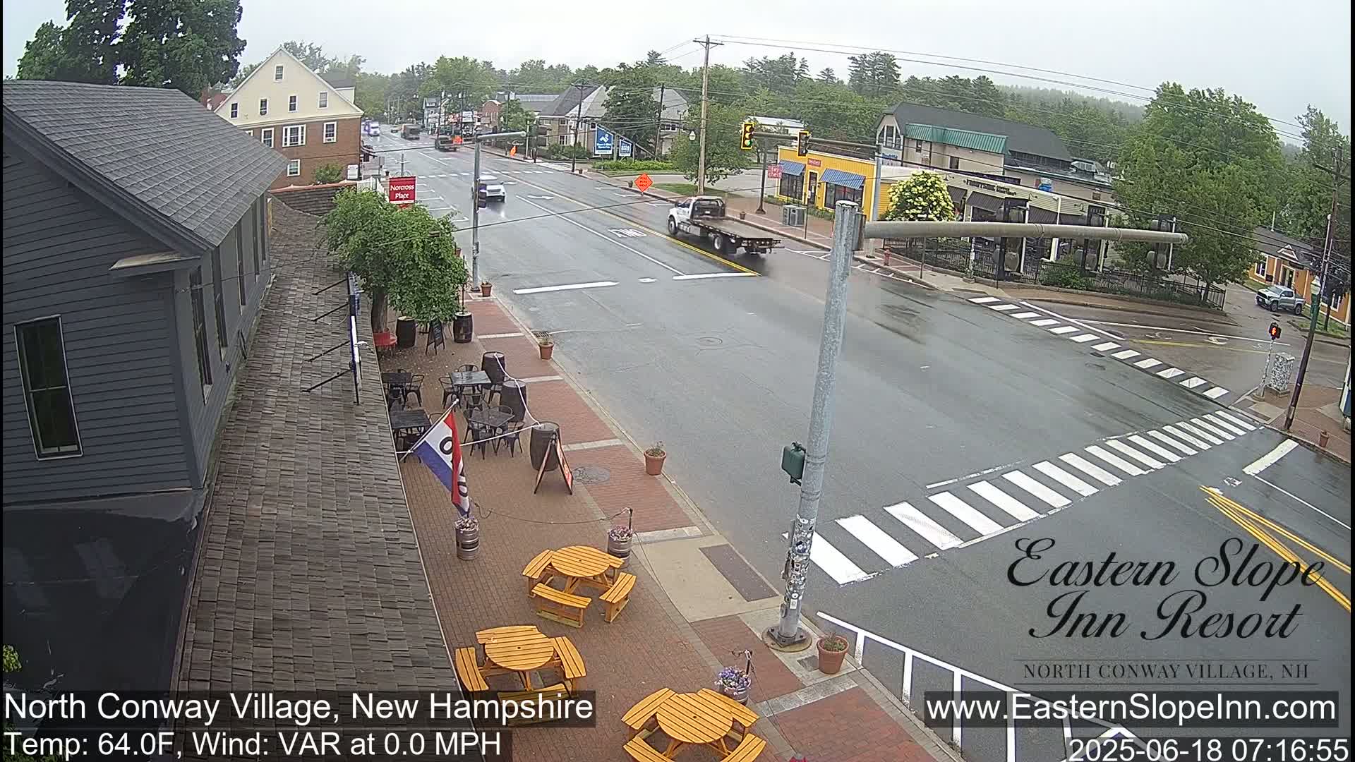 A wet street scene shows a town intersection with several buildings, outdoor seating, and sparse traffic under an overcast sky.