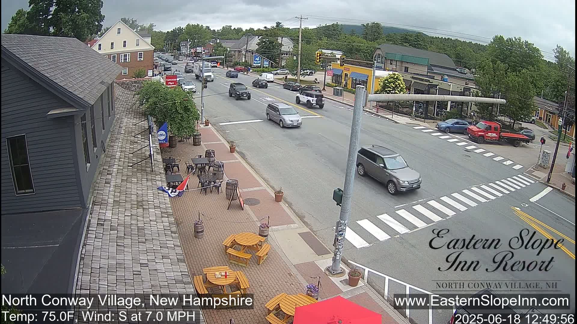A street scene on an overcast day shows a village street with cars, outdoor seating at a restaurant, and buildings under a partly cloudy sky.