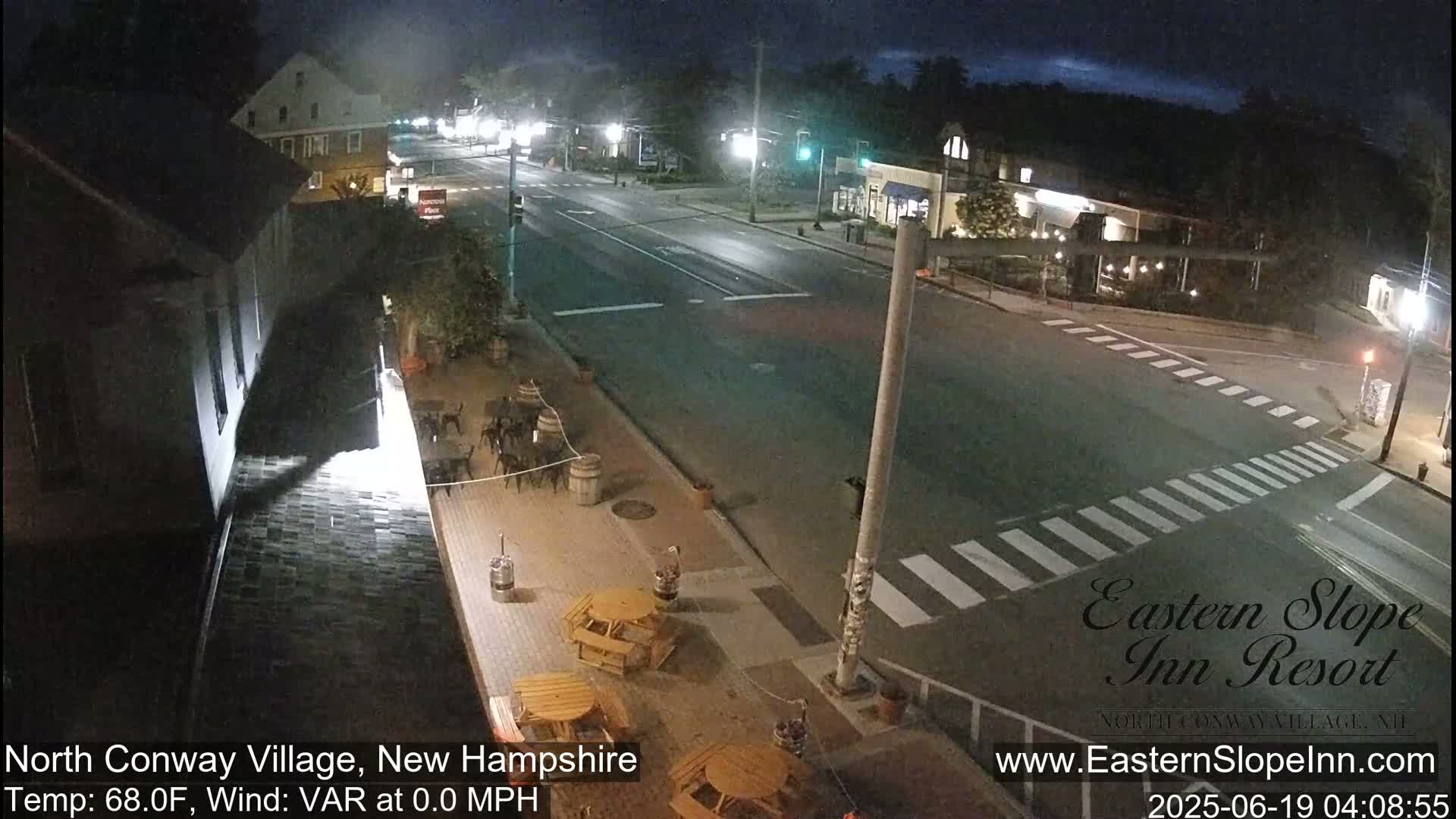 A nighttime street scene shows a mostly empty intersection with pedestrian crosswalks, buildings, and outdoor seating areas under a clear sky.